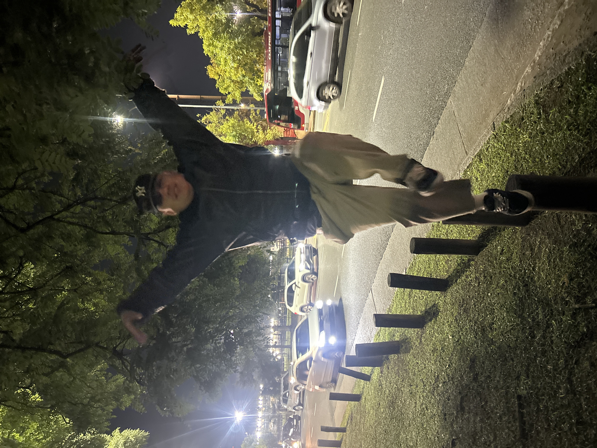 At night, a person is balancing on a low wooden barrier in what appears to be a public space, possibly a park or sidewalk in the city of Buenos Aires, Argentina. The person is wearing a baseball cap, jacket, and loose pants, with one foot on the barrier and arms outstretched. In the background, there is a street with cars parked and driving, illuminated by streetlights. Trees are present on both sides of the scene, and there is a red bus visible in the background. The wooden barrier is lined with grass.