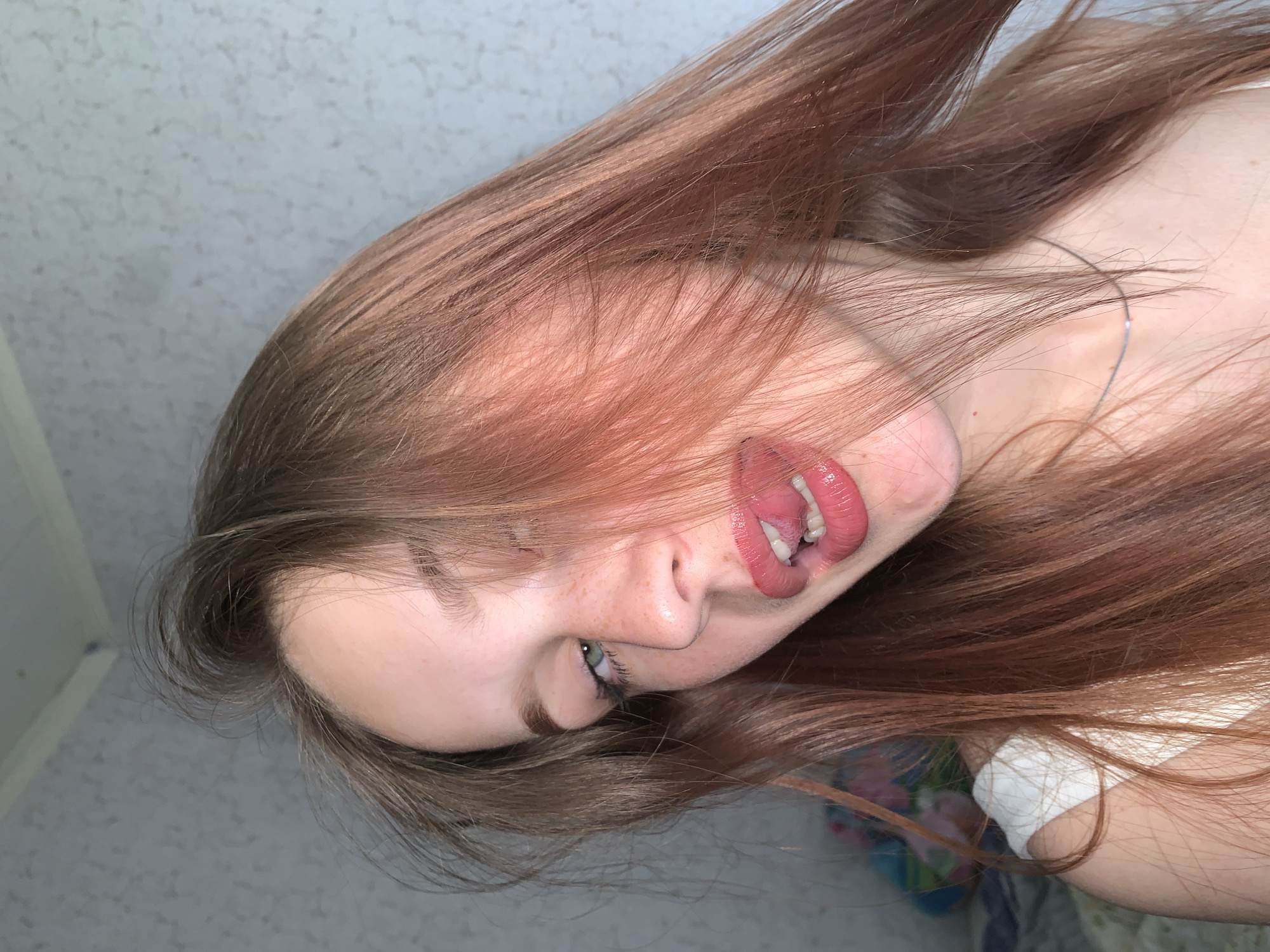 A close-up portrait captures a person, likely female, with long, flowing reddish-brown hair that partially obscures her face. The image is oriented diagonally, with the person's head tilted. She has fair skin, with a scattering of faint freckles visible on her nose and cheek.
Her left eye, with a light-colored iris (possibly blue or green), is visible and looks towards the viewer, framed by dark eyelashes. Her nose is slightly upturned. Her lips are coated with glossy pink or red lipstick. Her tongue is extended from her mouth and curled upwards, revealing a small, silver-colored stud piercing. Some white teeth are visible behind the tongue.
A thin, silver-colored necklace or chain is visible on her neck, peeking through her hair. A glimpse of a white garment, possibly a tank top or camisole strap, can be seen near her shoulder. The background is indistinct, primarily showing a light-colored, textured surface, likely an indoor wall or ceiling. In some areas, a blurry hint of colorful fabric, possibly a blanket or clothing, is visible behind her hair. The overall impression is a playful and expressive pose. No text is visible in the image. The location context is Фоки, Russia.