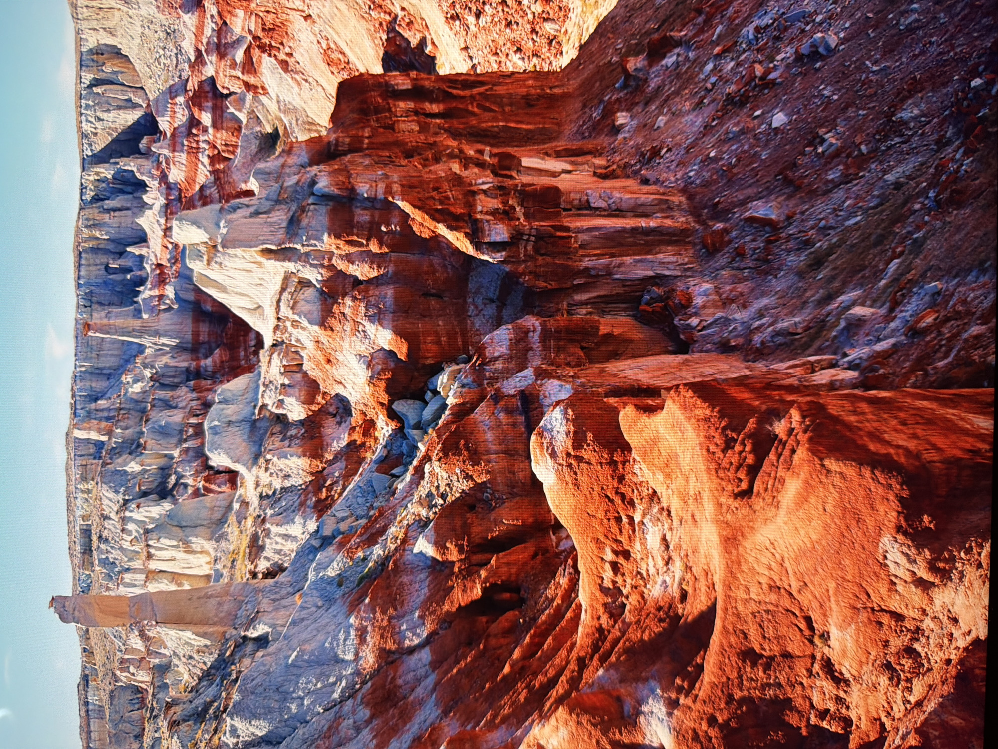 The image depicts a rugged, arid landscape characterized by towering rock formations and colorful stratified geological layers. The formations exhibit a variety of shapes, including spires, hoodoos, and cliffs, with colors ranging from deep red and orange to pale white and gray. The terrain appears eroded and weathered, suggesting a natural canyon or badlands environment. The sky above is a pale blue with some wispy clouds. There are no discernible man-made structures or human presence in the scene. The image does not provide any information or context that would place it within the city of Kyiv, Ukraine.