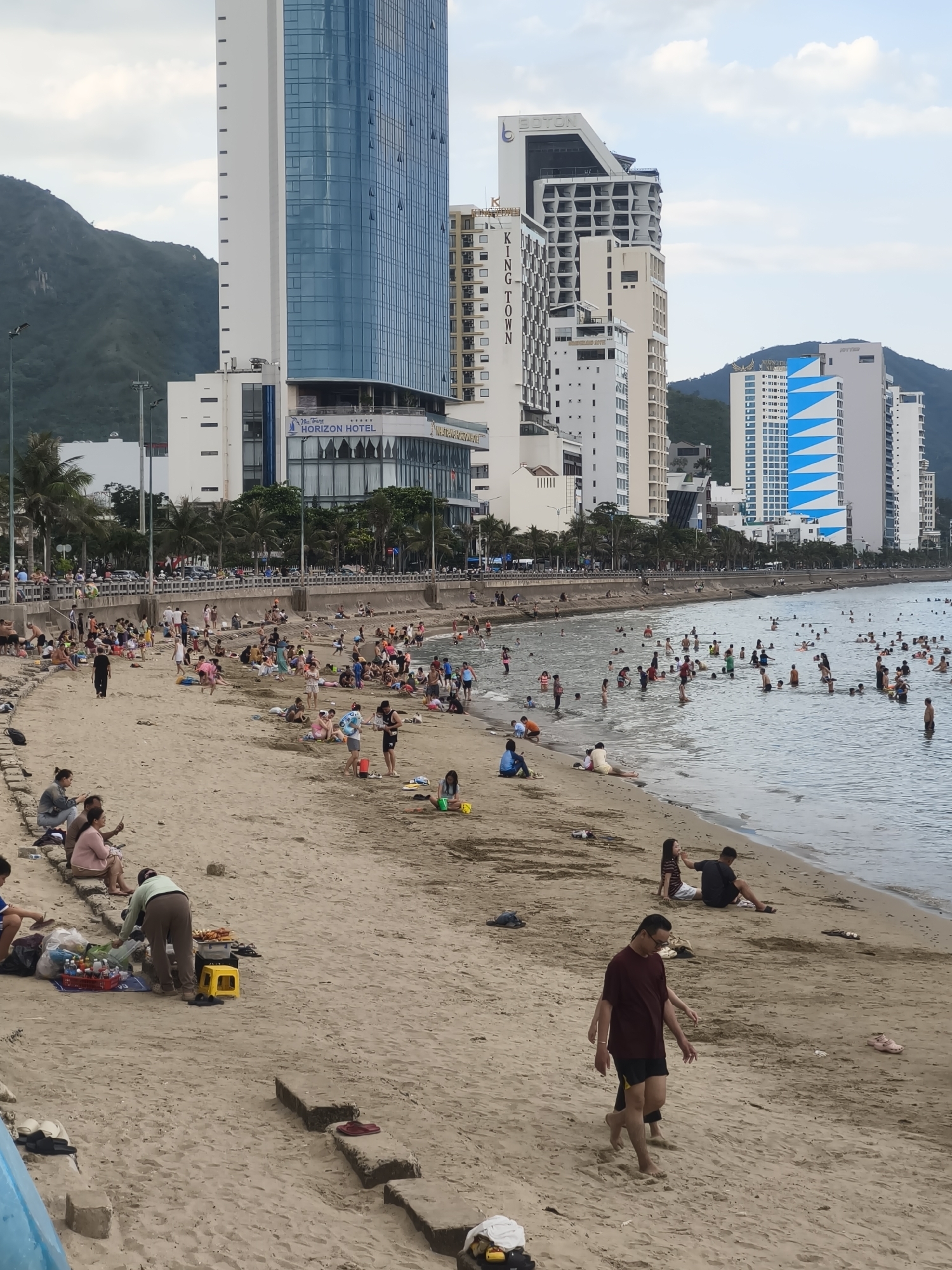This media file depicts a bustling beach scene in Nha Trang, Vietnam. The sandy shore is populated with numerous people engaging in various leisure activities. Some are wading and swimming in the calm, blue ocean, while others relax on the sand. Children can be seen playing, and groups of friends and families are gathered, enjoying the atmosphere.
The background is dominated by a striking line of modern, high-rise buildings, indicative of a coastal city resort. One prominent building features the text "THE HORIZON HOTEL" with a logo of a sailboat. Other buildings display geometric patterns and varied architectural styles. Lush green hills form a backdrop to the urban structures.
The time of day appears to be late afternoon, with soft, diffused light suggesting overcast or partly cloudy skies, typical of tropical weather. The overall mood is one of relaxed enjoyment and vacation vibes. The presence of beachgoers, the ocean, and the backdrop of hotels clearly establish this as a popular tourist destination. The sand is a light brown, with some scattered debris and a few instances of footwear visible. The water is a clear blue, with visible ripples and the shapes of people swimming. Several people are seen selling or preparing food on the beach, suggesting a vibrant local economy catering to tourists.