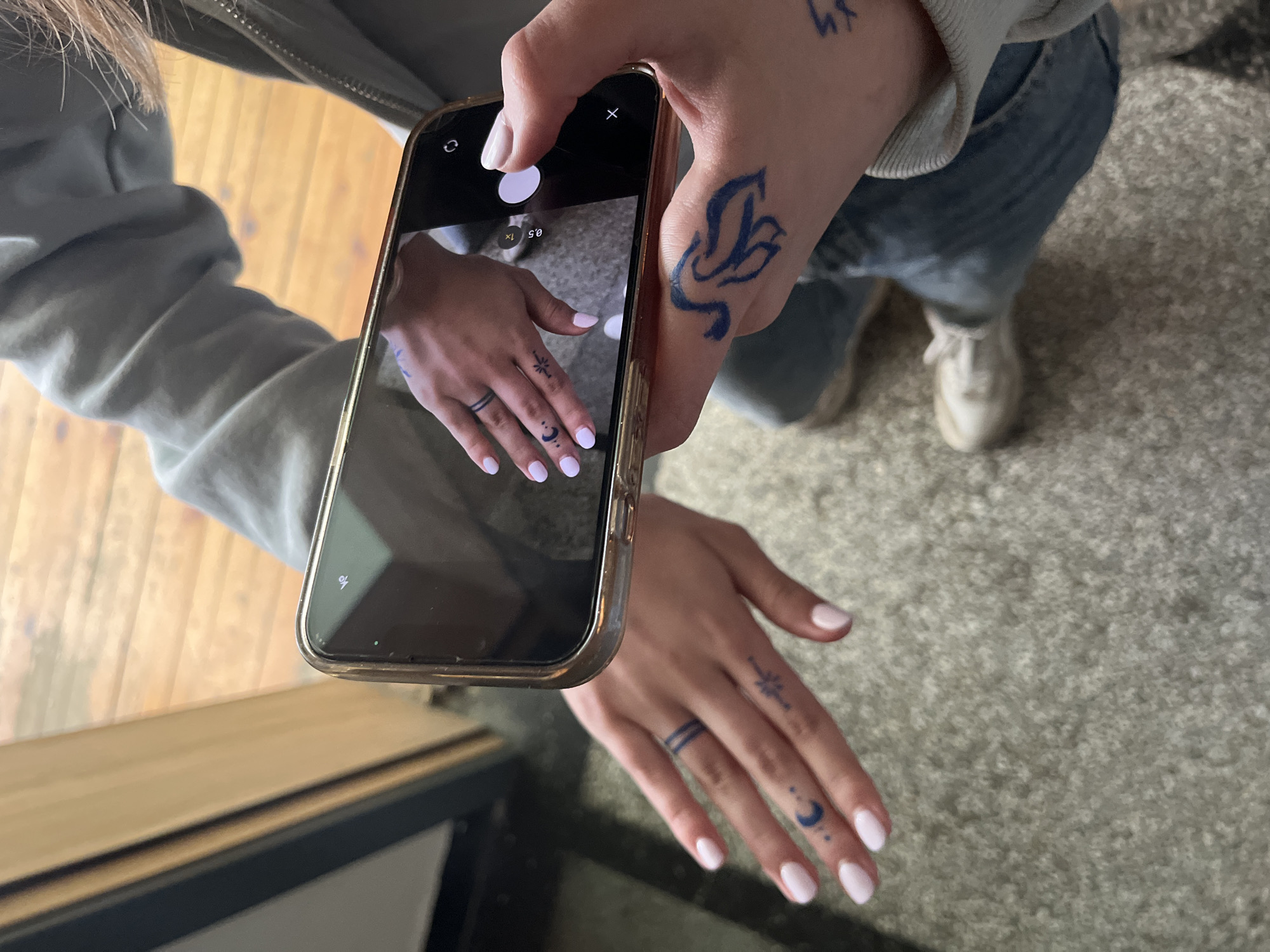 A person is holding a smartphone, taking a picture of their own hand, which is tattooed. The tattoos include a band around one finger, a moon and stars on another, and a floral design on the back of the hand. The person is wearing a grey jacket and jeans and standing on carpet. The location and city of Ruse, Bulgaria, cannot be verified from the visual information provided.
