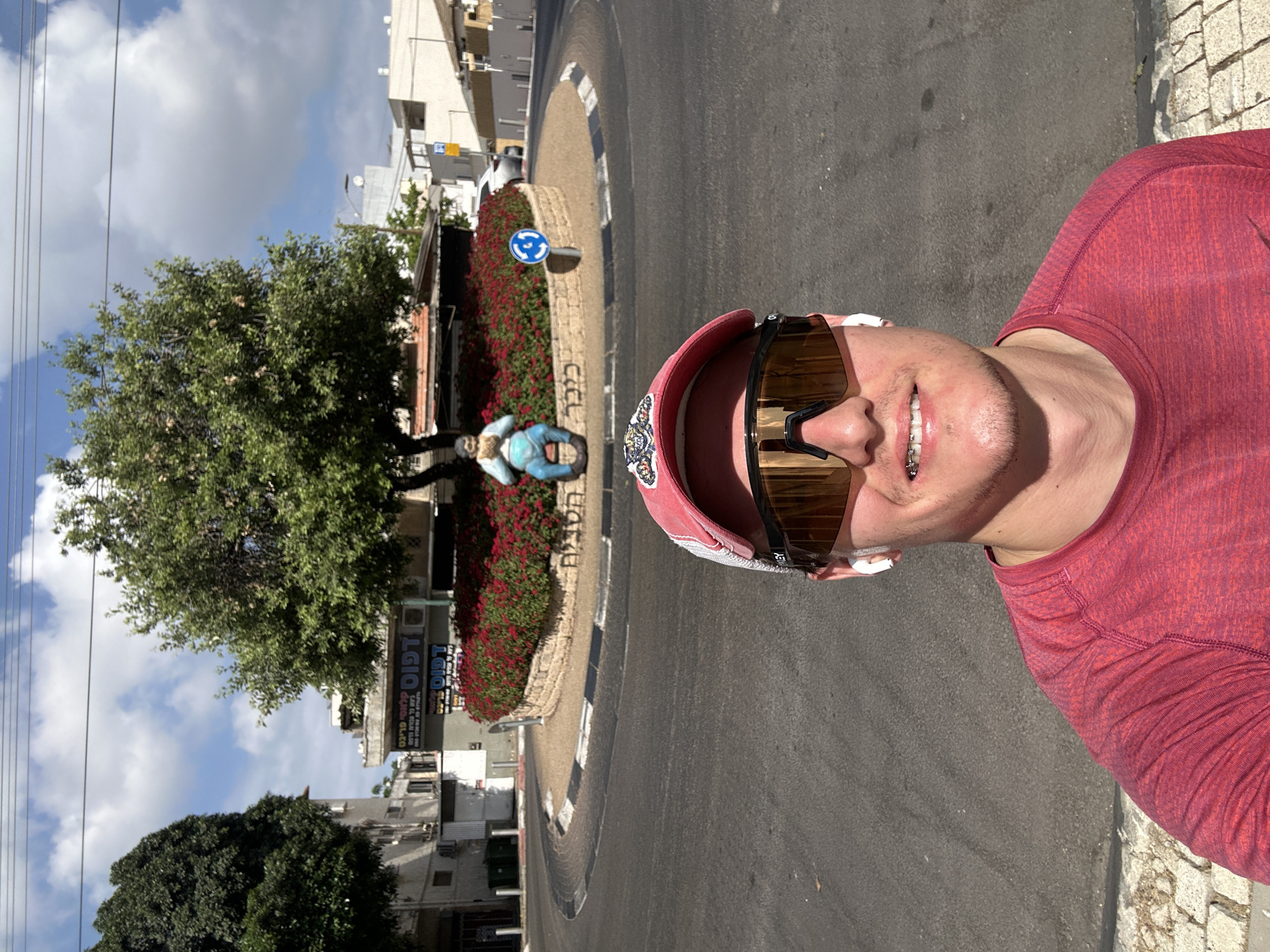 A man in a pink hat, sunglasses, and a red long-sleeved shirt is taking a selfie while standing in a street in Ramat Gan, Israel. Behind him is a roundabout with a tree, flowers, a statue of a person sitting, and a blue and white road sign. Buildings line the street in the background. The weather appears sunny with some clouds in the sky.