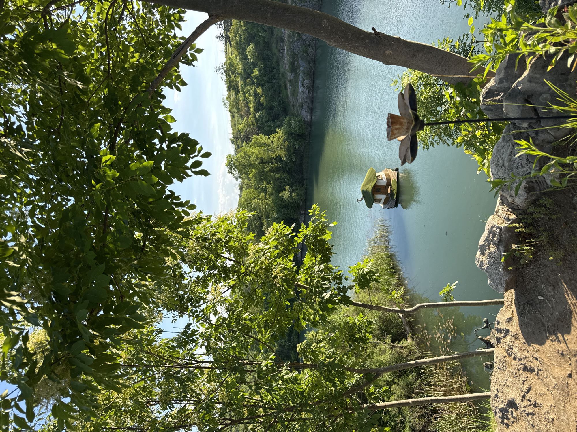 View from a high vantage point, possibly a quarry edge, overlooking a green lake. In the middle of the lake is a small, green-roofed building, possibly a birdhouse. In the right foreground, there is a rock outcrop and a metal flower sculpture. Trees and greenery frame the view. This is located in Lukovit, Bulgaria.