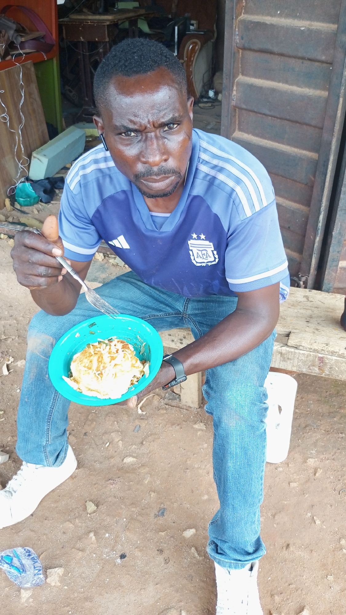 A medium shot depicts a man sitting outdoors, holding a bowl of food and a fork. The man, who appears to be of African descent, is wearing a blue Adidas t-shirt with the Argentinian soccer logo, blue jeans, and white sneakers. He has a dark complexion, short hair, and a slight frown on his face. He is wearing what appears to be a fitness tracker on his left wrist and an earpiece in his left ear.
He is holding a turquoise bowl containing a light-colored food item, potentially scrambled eggs or noodles, and a fork.
The setting appears to be an outdoor space in Barikin Sabon Gidan Kanat, Nigeria. The ground is dirt or reddish soil. The backdrop shows a wooden structure and other miscellaneous objects, indicating a somewhat cluttered environment. A white bucket sits near his right leg. The lighting suggests it is daytime.