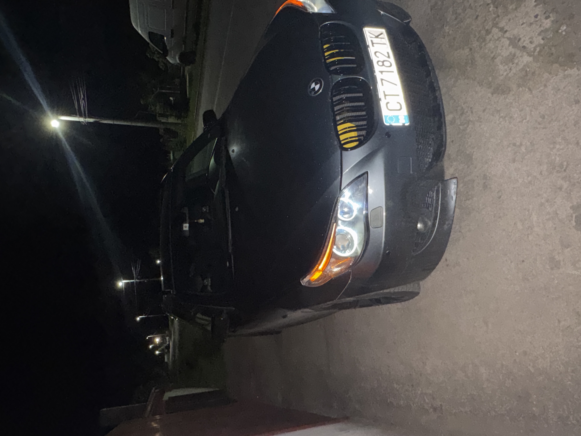 A black BMW car with the license plate CT 7182 TK is parked on a street in Sarnevo, Bulgaria, at night. The car is facing the right of the frame and is illuminated by its headlights and nearby streetlights. The car door is open. The surrounding environment includes other parked vehicles, sidewalks, and buildings visible under the dim lighting conditions.