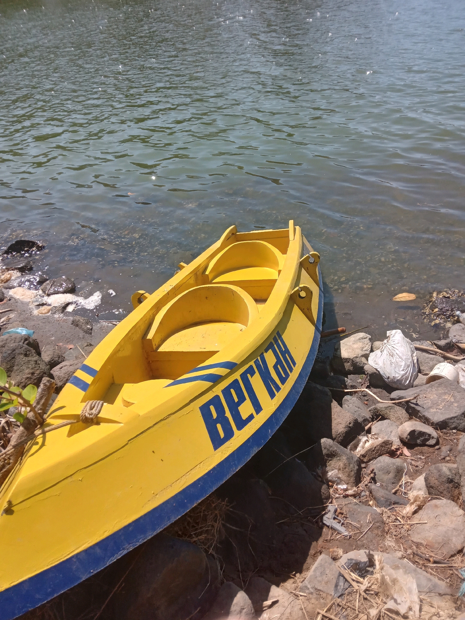 A bright yellow kayak with blue trim rests on a rocky shore beside a body of water that reflects the sunlight. The kayak has the word "BERKAH" written in blue letters on its side. The water appears calm with ripples on the surface. The shore is littered with rocks, dried leaves, and some discarded plastic bags and bottles, indicating a potentially unkempt or natural environment. The scene is lit by natural daylight, casting shadows on the kayak and the shore. The overall impression is a quiet, possibly a scenic, location that could be near the coast or a large lake in Wates, Indonesia, given the context.