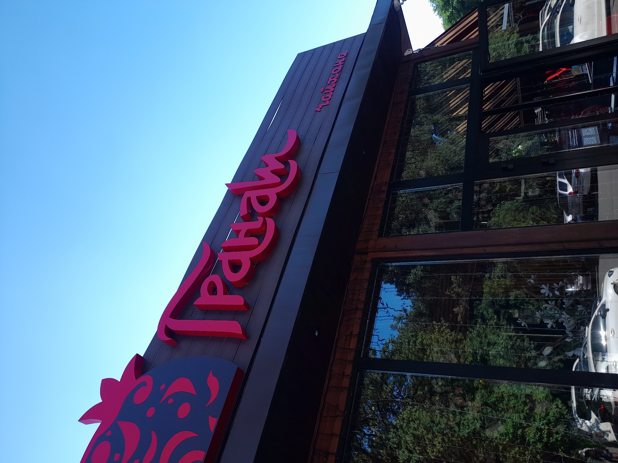 The image shows the exterior of a building with a prominent red sign that reads "ГРАНАМ" in large, stylized Cyrillic letters. To the left of this sign is a graphic of a stylized strawberry. Below the main sign, in smaller red Cyrillic script, is the word "чайхана" (chaikhana), indicating a tea house. The building has large glass windows reflecting trees and the sky. The architecture suggests a modern establishment with wooden accents. The sky is clear and blue. The location is identified as Vladivostok, Russia.