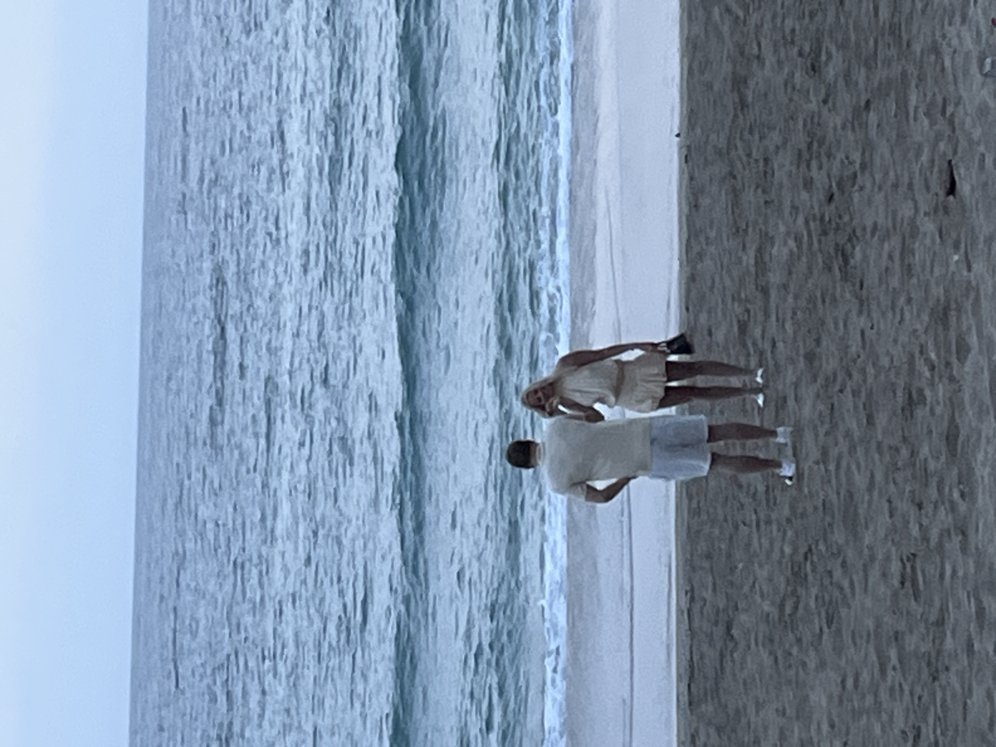 A medium shot captures a man and a woman standing on a sandy beach adjacent to an ocean. They are both facing the ocean, with the man slightly ahead of the woman. The ocean waves are visible in the background, and the sky appears overcast. The man is wearing a light-colored short-sleeved shirt and shorts, while the woman is wearing a light-colored dress. They appear to be walking together near the shoreline. This photograph was taken in Alanya, Turkey.