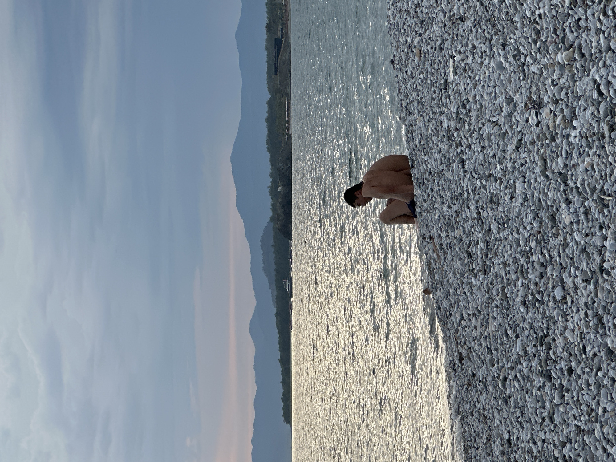 A man sits on the rocky beach of Butterfly Valley in Fethiye, Turkey. The beach is covered in small, light-colored rocks. Water glistens in the center of the frame. Beyond the water, mountains are visible, and the sky is light blue.