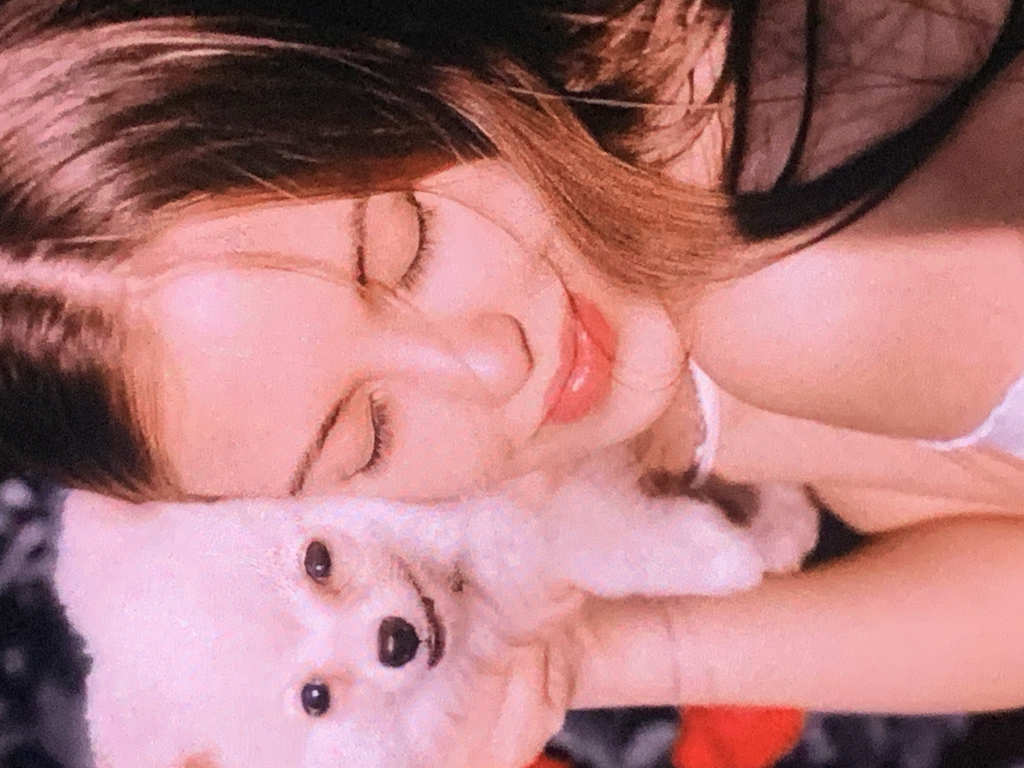 The image is a close-up, slightly blurry, and overexposed shot of a young woman holding a small white dog. The woman's eyes are closed, and her head is tilted slightly. She has long, dark brown hair. The dog is fluffy and white with dark eyes and a small black nose. The woman appears to be wearing a white top and black underwear. The background is dark and indistinct, with some red shapes that might be flowers. The overall color palette is warm, with strong pink and orange tones. There is no discernible location in Sevastopol, Ukraine, or any other city.
