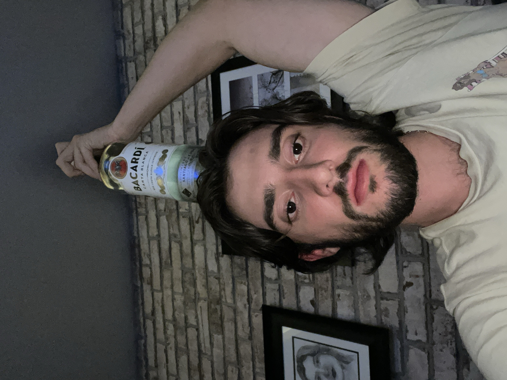 A man is lying down, holding a bottle of Bacardi Carta Blanca rum on his head. The background includes a brick wall and two framed pictures. The location is likely indoors, but specific details of the environment, such as the city of Huixquilucan de Degollado, Mexico, cannot be determined from this image alone.