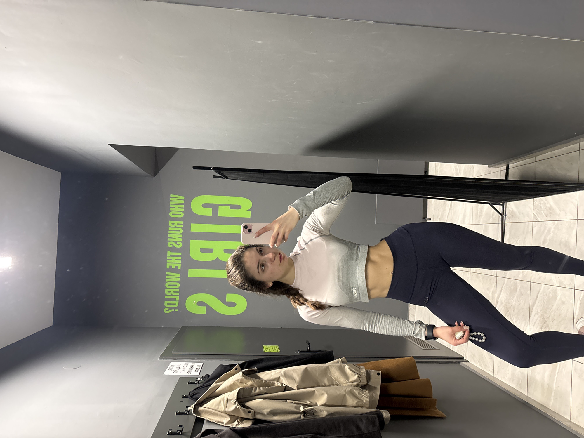 A woman in workout clothes holds a phone, taking a selfie in a gym locker room. The locker room has gray walls with the words "GIRLS, WHO RUN THE WORLD?" written in green. There are coats and jackets hanging in the locker room. The city and country are Unknown.