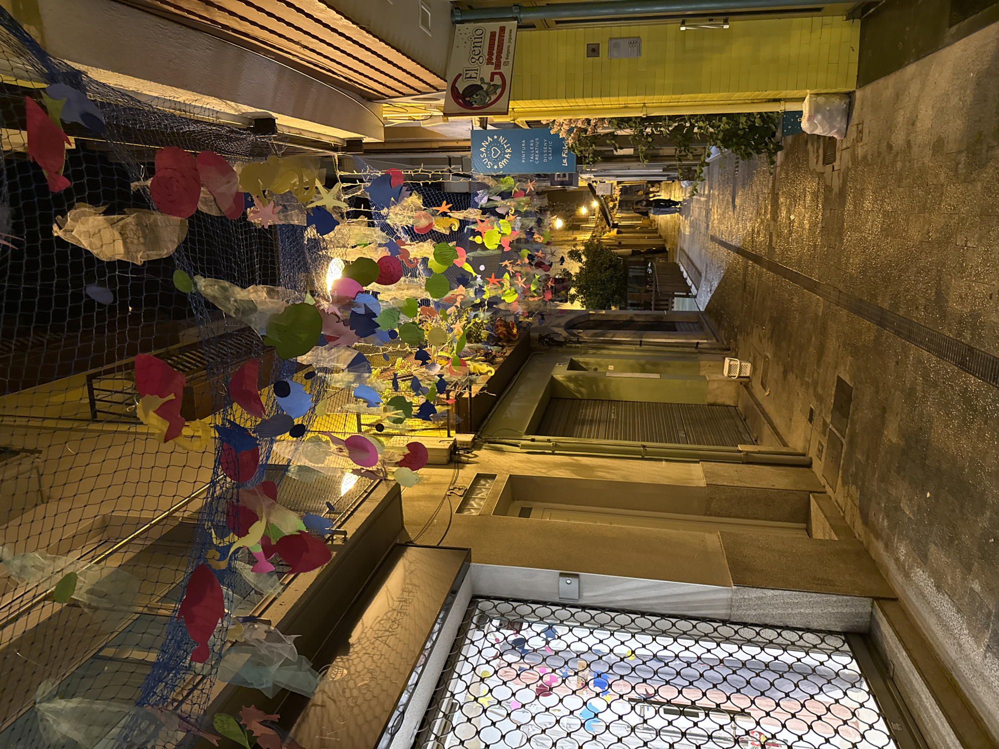 The image depicts a narrow street in Blanes, Spain, decorated with colorful paper cutouts of marine life and netting strung across the street. The street is paved with wet cobblestones, reflecting the warm glow of streetlights and building lights, suggesting it may have recently rained or is damp. Buildings line both sides of the street, with storefronts visible. One storefront on the left has a metal security gate lowered, revealing a display of similar colorful decorations behind it. On the right, a brightly painted yellow building features a sign for "El Genio" and another blue sign with text that includes "SUSANA SMARTIN." Two people are walking away from the viewer down the street. The overall scene conveys a festive atmosphere, likely part of a local celebration or event.