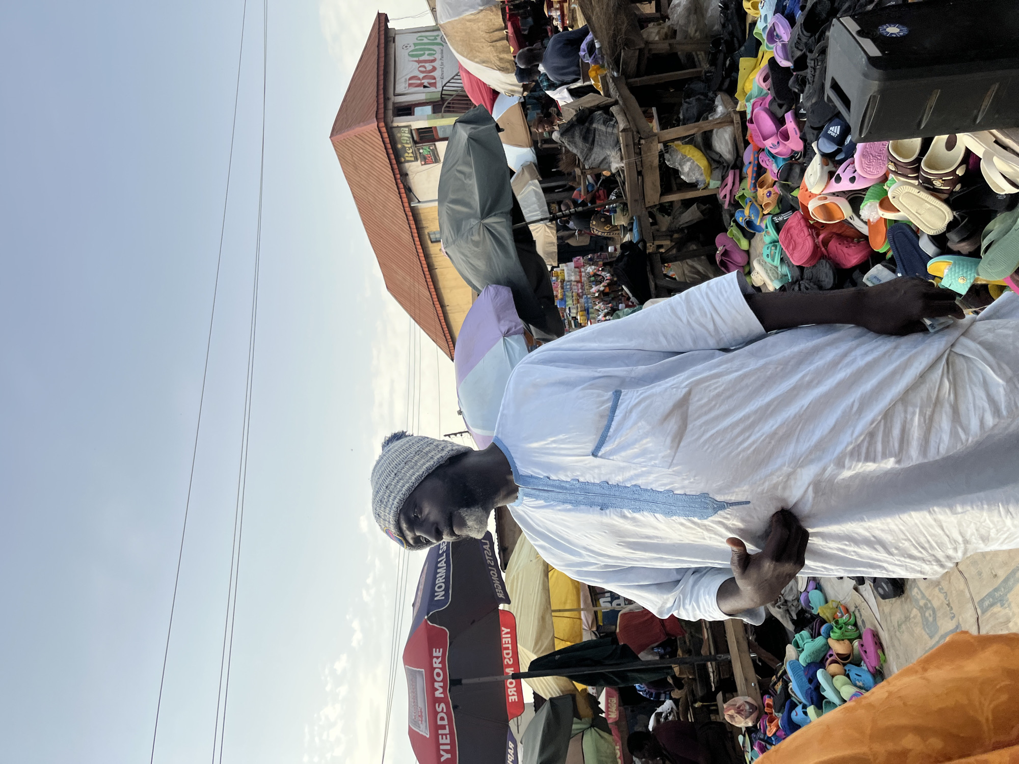 A man wearing a white garment and a grey knitted hat stands in a market. He is looking downwards with a neutral expression. In front of him, a large pile of colorful flip-flops and sandals is displayed for sale. Behind the man, stalls are visible, displaying more merchandise and awnings. The scene appears to be outdoors in a bustling marketplace. The lighting suggests it is daytime, possibly late afternoon, with a clear sky overhead. The overall setting is a vibrant and busy market environment. A sign above some of the stalls reads "Bet9ja."
