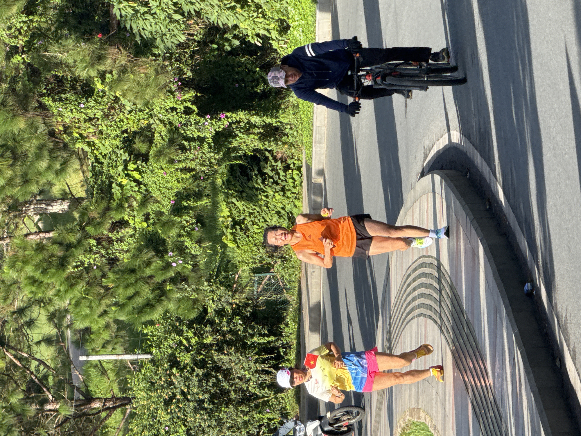 In Phường Xuân Hương - Đà Lạt, Vietnam, under a bright sky casting long shadows, three individuals are engaged in outdoor activity on a paved pathway and adjacent road. On the left, a woman wearing a white cap, a brightly colored yellow, blue, and pink athletic shirt, and pink shorts is running. She has a white armband with red and yellow markings. To her right, a woman in an orange sleeveless athletic shirt and black shorts is also running, holding a small yellow object in her right hand. Further to the right, a person wearing a dark blue hoodie, black pants, black gloves, and a camouflage-patterned baseball cap is riding a bicycle. The bicycle is dark colored with red accents on the wheels. The environment is lush and green, with dense foliage and trees lining the background, suggesting a park or scenic route. A motorcycle is partially visible on the far left. The paved path features a distinctive patterned section with curved lines, and the road is asphalt.