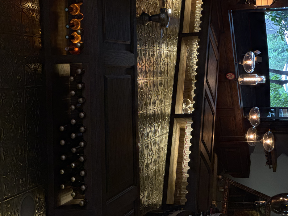This is an interior, eye-level photograph of a bar or restaurant in Mexico City, Mexico. The scene is dominated by dark wood paneling and an ornate, gold-toned embossed metallic ceiling. Above, a long, recessed dark wooden shelf holds approximately 25 dark-colored bottles horizontally, consistent with wine storage. To its right, a smaller, illuminated cubby displays four upright, amber-liquid-filled bottles. Directly below, multiple rows of clear, inverted stemware, including wine and shot glasses, are suspended from wooden racks. A metallic ceiling fan with an integrated light fixture is visible on the upper right. Five hanging pendant lights with clear glass globes and visible incandescent filaments illuminate the mid-ground. A red and white circular "No Smoking" sign is affixed to a central wooden support. A portion of an ornate, gilded frame is visible on the far left. Through a window or doorway on the lower right, green outdoor foliage is discernible. No individuals or active events are present.