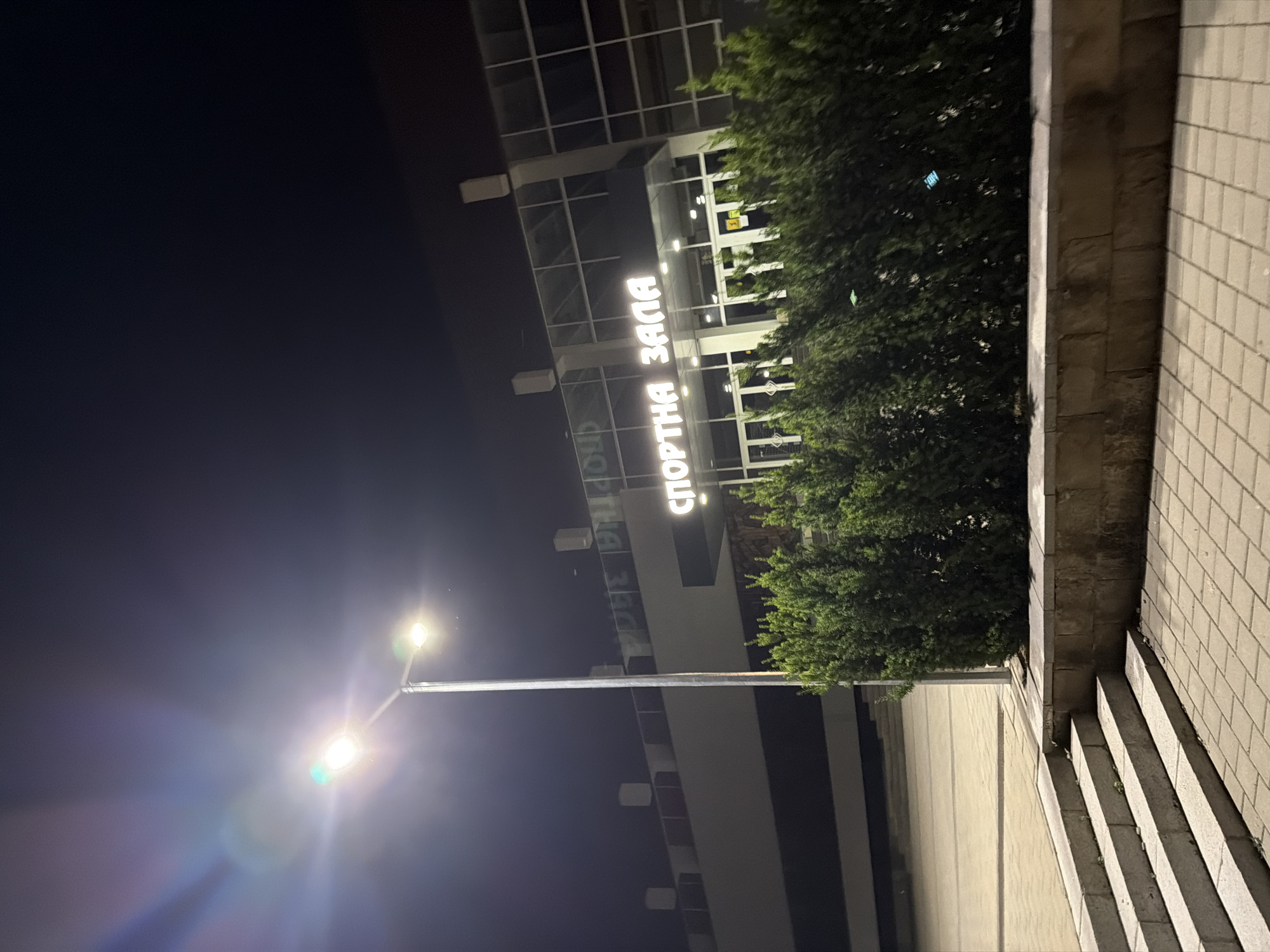 The image shows the exterior of a sports hall at night in Pravets, Bulgaria. The hall has large glass windows and illuminated signage that reads "СПОРТНА ЗАЛА" (Sports Hall). A tall street lamp with two lights illuminates the foreground, casting lens flares. In front of the building is a row of manicured evergreen bushes, and to the left, there are stone steps leading up to a paved area. The overall environment suggests an outdoor public space at night.