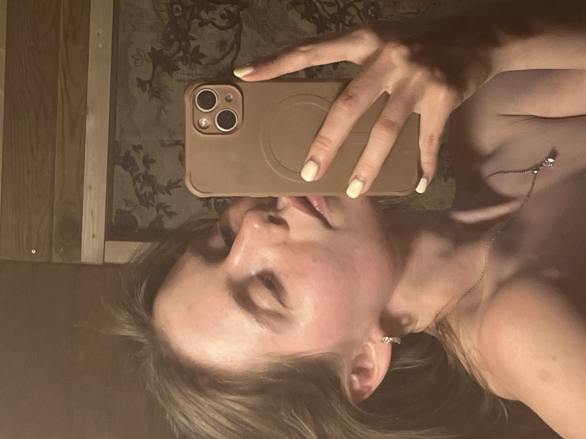A woman with light skin and blond hair is holding a tan-colored iPhone in front of her face, appearing to take a selfie. She is wearing a delicate silver necklace and an earring in her left ear. Her nails are painted a light yellow color. The background appears to be a mirror reflecting a wall with patterned wallpaper and a wooden frame. There is no indication of this image being taken in the city of Chaikovsky, Russia.