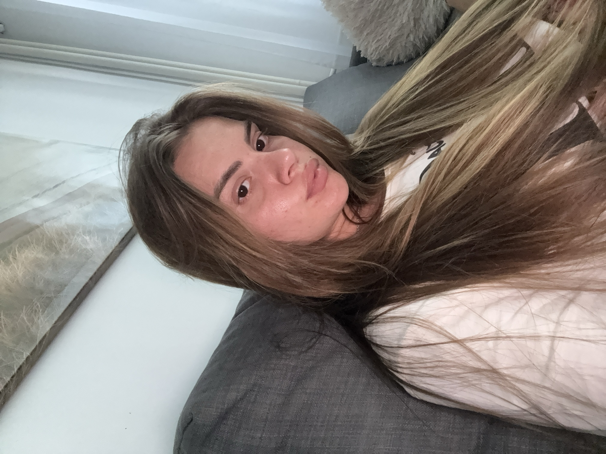 In a domestic indoor setting, a young woman with long, blonde-brown hair is seated on a dark gray couch. She is looking directly at the camera, with her body angled slightly to the left. She is wearing a white t-shirt. The background includes a white wall, sheer white curtains to the right, and what appears to be artwork on the wall above and behind her. There are no discernible geographical markers or indications that would place the scene in Plovdiv, Bulgaria.