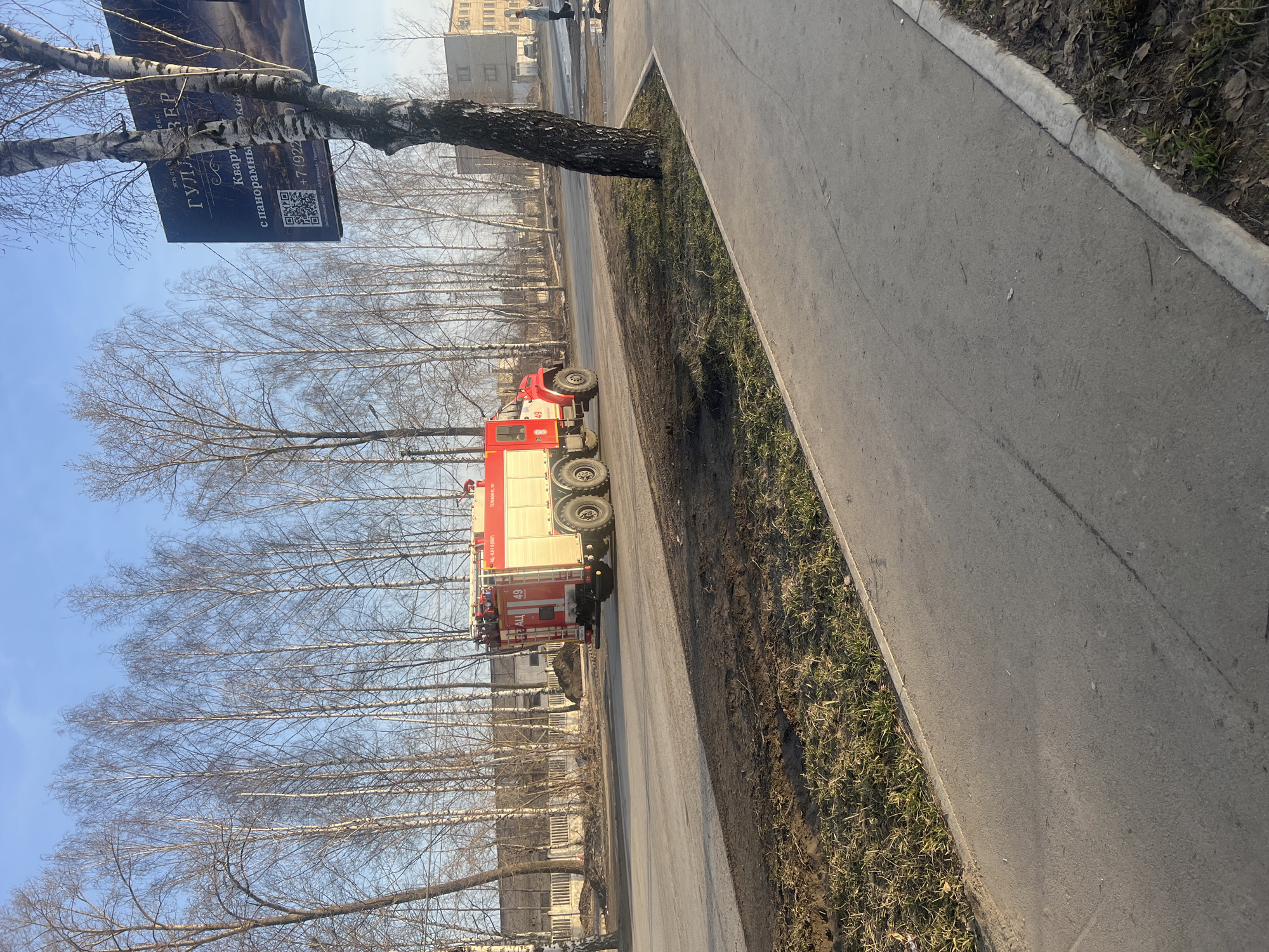A fire truck is located on a street, with a tree along the sidewalk and a commercial billboard. Based on the markings on the fire truck, this image may be located in the city of Chaikovsky, Russia.