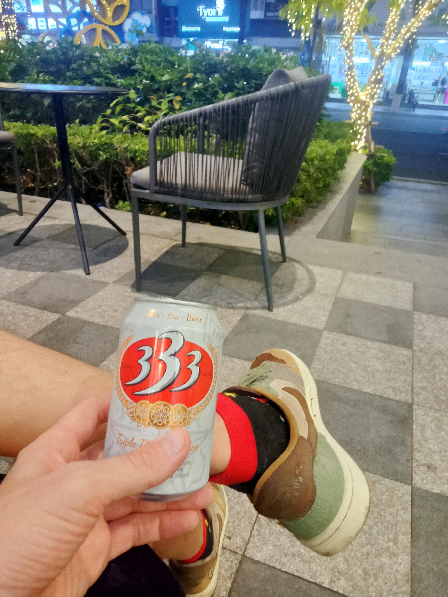 A person is sitting on a tiled surface outdoors, holding a can of "333" beer. The person is wearing a pair of camouflage sneakers with red socks. Behind the person is a small black table and a dark gray chair with a light gray cushion. The chair and table are situated near a raised garden bed filled with green bushes. In the background, there are buildings and trees decorated with string lights. A business sign with the name "Tymi?" is visible, indicating a possible business establishment in the background. This scene is likely occurring in the city of Nha Trang Ward, Vietnam, based on the "333" beer.