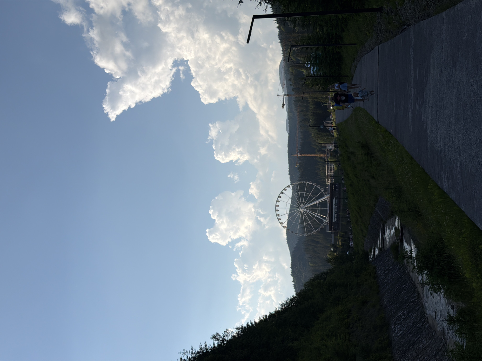 This image captures a scenic view in Polianytsia, Ukraine, featuring a large Ferris wheel prominently in the background. The scene is dominated by a clear blue sky with scattered white clouds, characteristic of a bright day. In the foreground, a paved pathway winds through a grassy area, with a small, shallow stream flowing alongside it. A steep, tree-covered slope rises on the left side of the pathway, contrasting with the open landscape on the right.
Several people are visible walking along the pathway. A family, including two adults and two children, are the most prominent figures, with the younger child running ahead. Further down the path, other individuals are also walking. The Ferris wheel, a significant attraction, stands on a hill surrounded by a dense forest. Two construction cranes are also visible in the middle ground, suggesting ongoing development or maintenance in the area. The overall atmosphere is tranquil and picturesque, with a sense of a popular tourist destination.