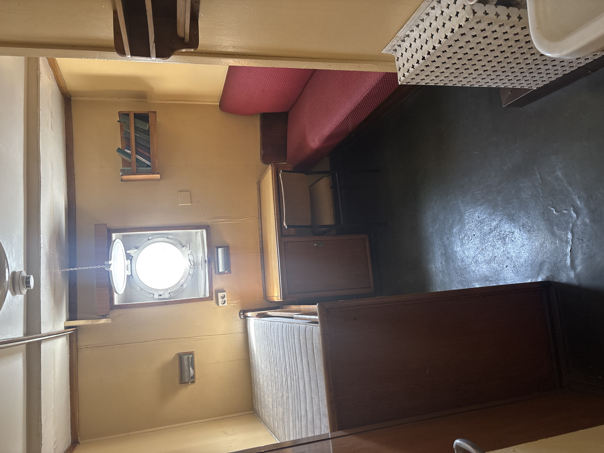 The image shows a small, windowed room with yellow walls. On the left, a single bed with a grey mattress is visible, partially enclosed by a wooden frame. A wooden desk with a drawer and a metal-framed chair with a beige seat and back are placed in the center of the room. Above the desk, a circular porthole window with a metal frame is set into the wall, letting in bright light. To the right of the desk, a long, red upholstered bench seat is against the wall. Above the porthole window, a small wooden shelf holds a few books. To the right of the bench seat, a white radiator cover with a decorative pattern is visible, and next to it, the edge of a white sink. The floor is dark and appears to be concrete. The room has a simple, functional aesthetic, likely a cabin on a ship or a similarly compact living space.