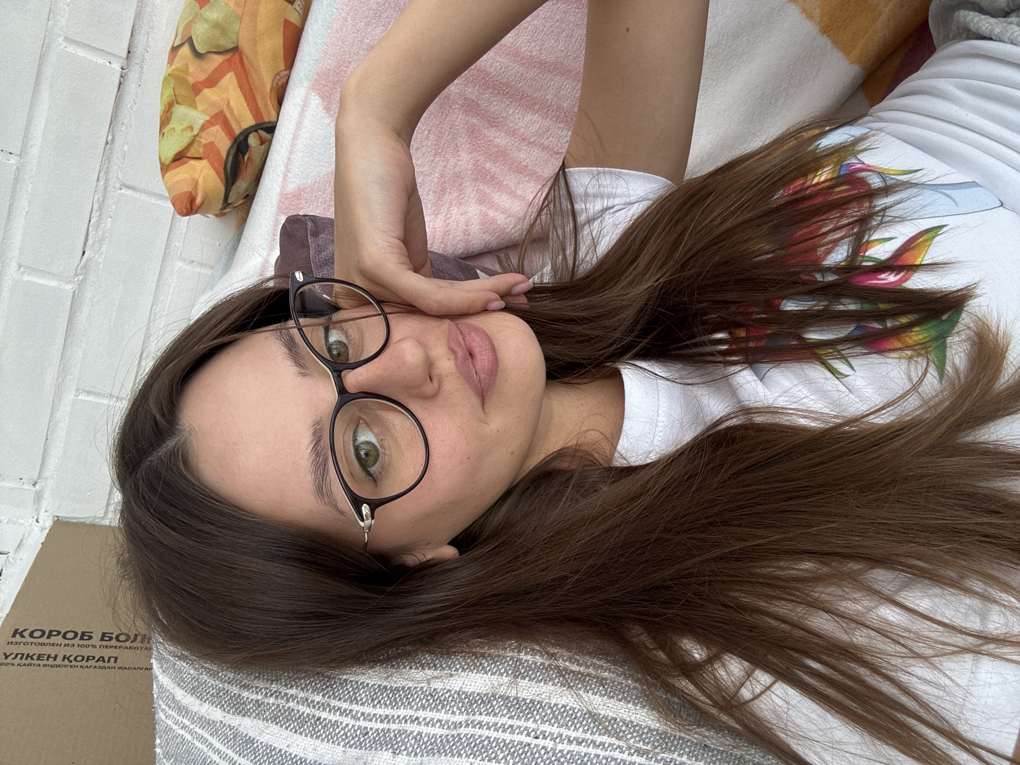 A young woman with long, brown hair and green eyes is captured in a close-up selfie. She is wearing glasses with dark frames and a white t-shirt with a colorful, abstract graphic on it. Her left hand is resting on her cheek, and she has light pink nail polish on. She appears to be lying down on a textured surface, possibly a couch or bed, with striped pillows and a pillow with an orange and yellow abstract pattern behind her. The background shows a white brick wall. The scene suggests a casual, indoor setting. There is a cardboard box with text in Cyrillic script visible in the upper left corner, partially obscured by her hair. The text on the box includes "КОРОБОК БОЛЬ", "100% КАРТОНА", and "ИЗГОТОВЛЕН ИЗ 100% ПЕРЕРАБОТ". Given the Cyrillic text and the location of the photograph being in Krasnoyarsk, Russia, it is likely an indoor setting within the city.