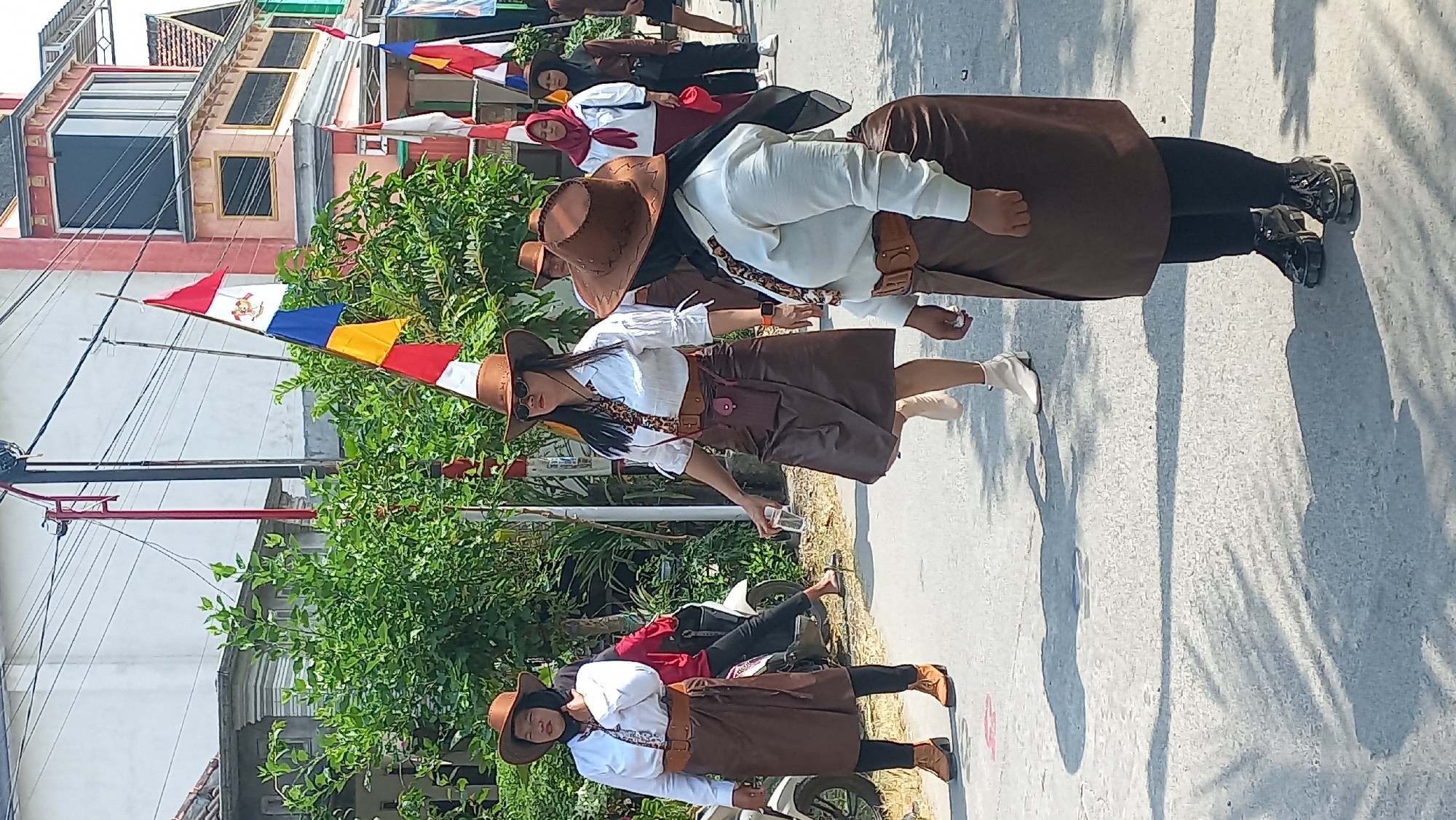 In Kedungmegarih, Indonesia, a group of women, dressed in matching brown hats, white shirts, and brown skirts, are participating in a parade or procession along a paved street. The women are walking in single file, some holding cups. A motorcycle is parked on the left side of the street, and several Indonesian flags are visible in the background, along with buildings and lush green trees. The sun is casting shadows on the street, indicating a daytime scene.