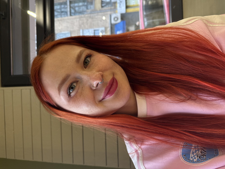 A close-up, chest-up portrait features a female individual indoors, consistent with an urban environment in Rostov-on-Don, Russia. The subject has long, bright red hair, fair skin, and visible freckles. She has blue-green eyes, wears black eyeliner and dark pink lipstick, and a silver septum ring in her nose. She is looking directly at the camera with a subtle smile. She is dressed in a light pink long-sleeved top with a white collar band and a light blue patch on the left chest area, featuring a checkered flag and text including "WORLDWIDE" and "CAPITOL". The immediate indoor background to the left consists of light-colored horizontal wall paneling. Through a window in the upper right, an outdoor urban scene is discernible, revealing a multi-story building, a partial blue sign, a white exterior air conditioning unit, and the upper section of a red vehicle, indicating a street beyond.