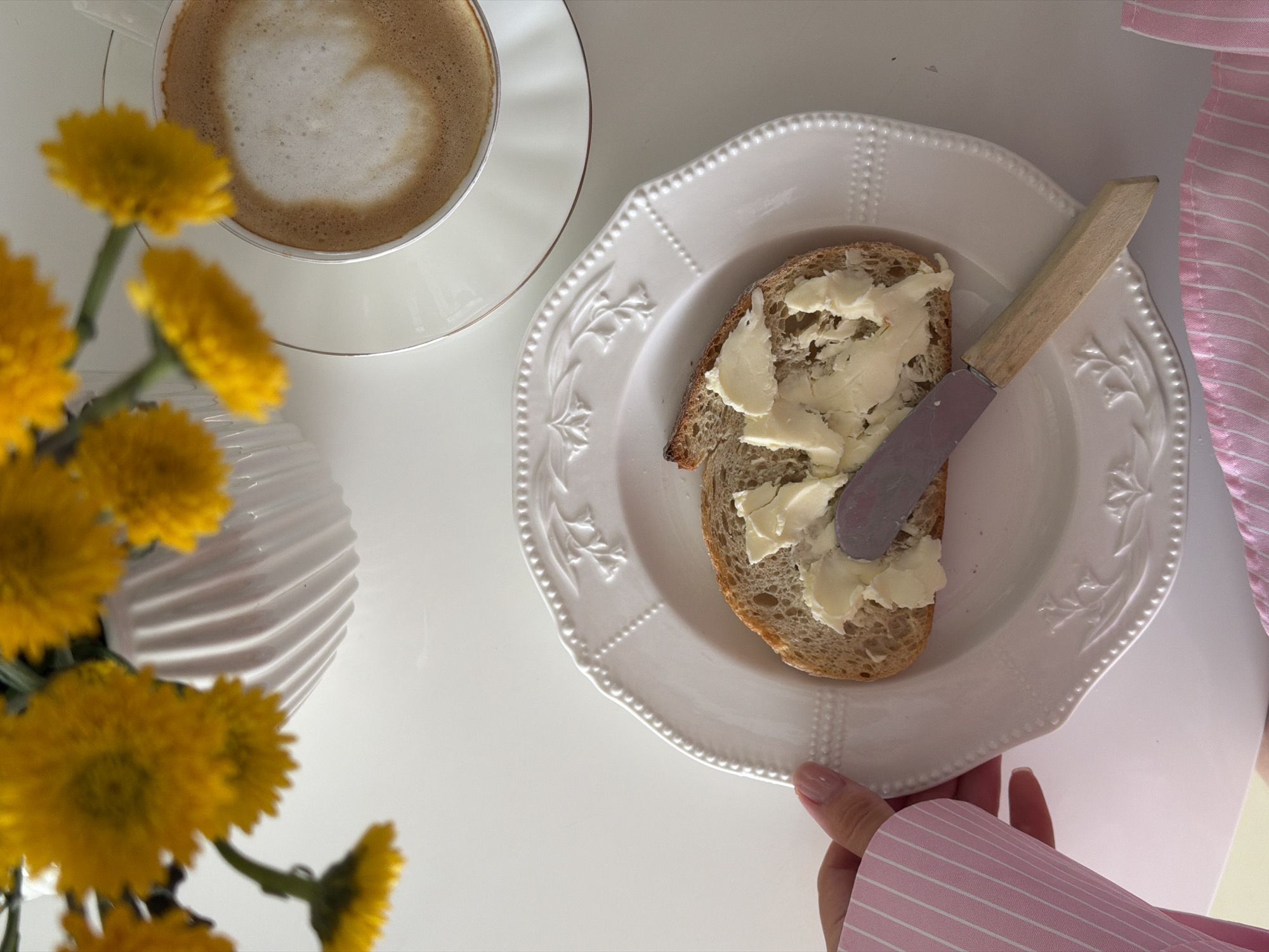 A high-angle shot shows a white table with breakfast or a light meal. A cup of cappuccino sits on a saucer at the top left, near a vase of yellow flowers. A white plate with a slice of bread covered with butter and a knife rests in the center. A person wearing a pink striped shirt holds the plate from the bottom right. The location is unknown, and there is no indication this image was taken in Khimki, Russia.