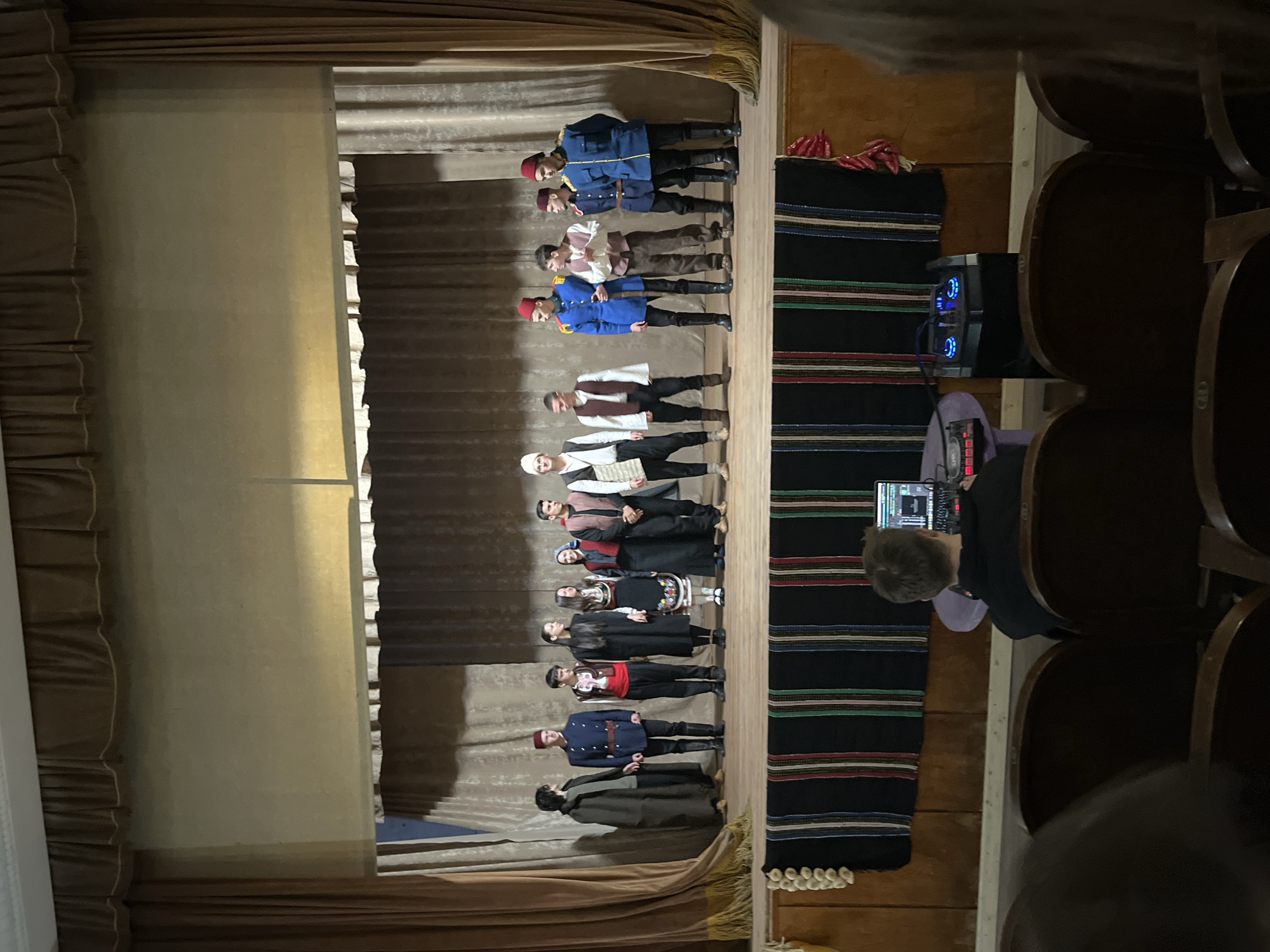 A wide-angle shot shows a theatrical performance in what might be the town hall of Voiniagovo, Bulgaria. A group of roughly a dozen people is standing on stage wearing traditional Bulgarian costumes. The stage has a simple setup with beige curtains on either side and at the top.
To the right of the stage, a black carpet with colorful stripes is seen. A person is sitting in the audience, operating sound equipment. Rows of wooden seats can be seen in the foreground. The location is indicated by a sticker with the town's name on the wall.