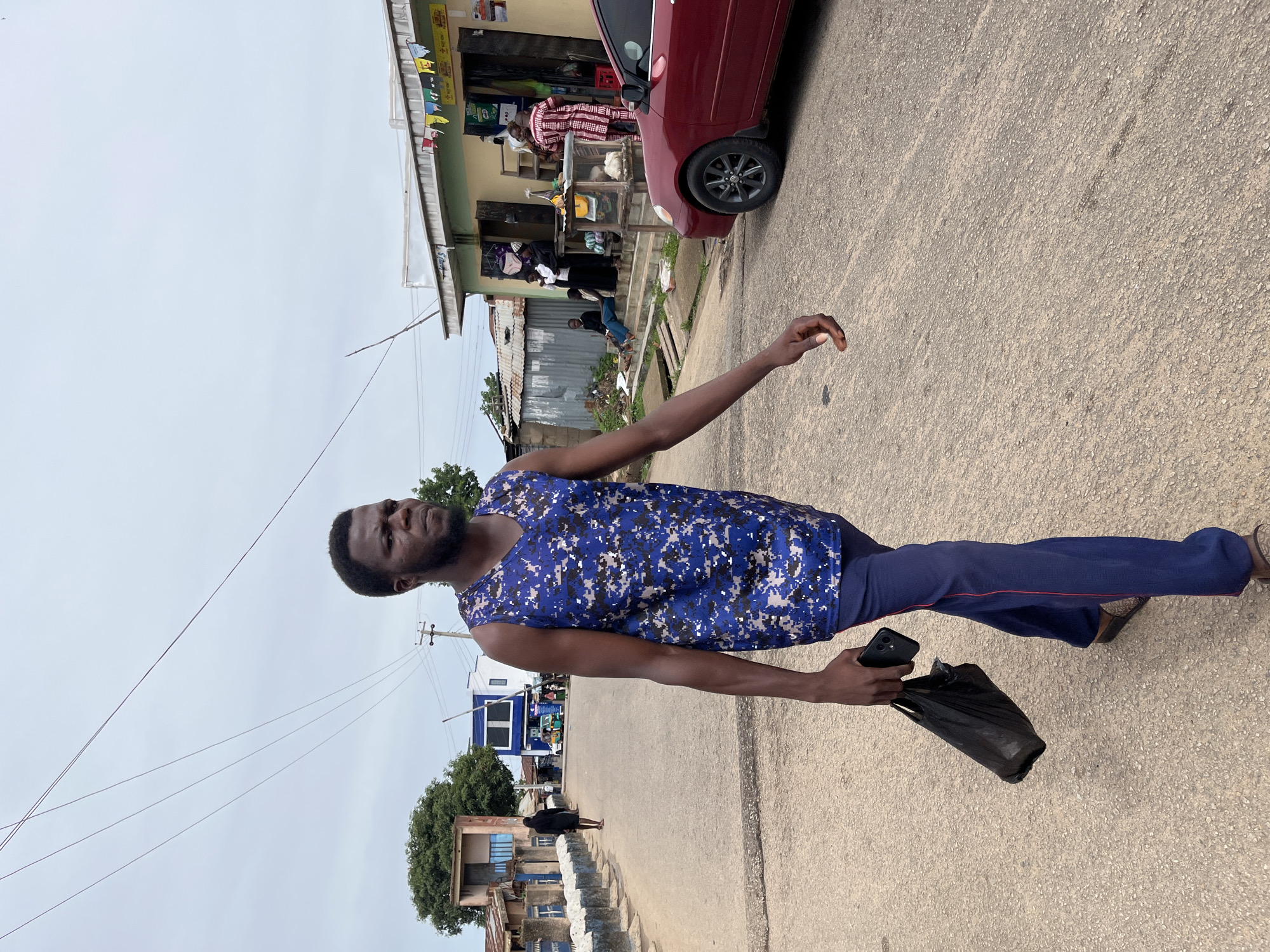 A full-shot image, rotated 90 degrees clockwise, captures a man standing on a paved road under an overcast sky in what appears to be a local street scene in Jos, Nigeria.
The central subject is a man with dark skin, short hair, and a short beard. He is wearing a blue and white digital camouflage pattern sleeveless top and dark blue trousers with a thin red stripe running down the side. He holds a black smartphone and a black plastic bag in his left hand, while his right arm is slightly extended outwards with an open hand. He looks upwards and to his left, seemingly at something outside the frame.
In the background, to the man's right, a dark red sedan is parked partially on the road next to a single-story building that appears to be a shop or market stall. A man in a red and white striped traditional outfit is visible near the shop entrance, with other people also present around the building. Colorful, small banners are strung across the shop's facade, but any text on them is unreadable.
To the man's left, further down the road, another person in dark clothing walks away. The street is lined with simple, low-rise buildings, including a light-colored structure with a blue accent. Utility poles and overhead power lines traverse the grey, cloudy sky. The ground is a rough, light-colored paved road. The scene depicts a typical daily moment in an urban or semi-urban Nigerian setting. No distinct legible text is visible in the image.