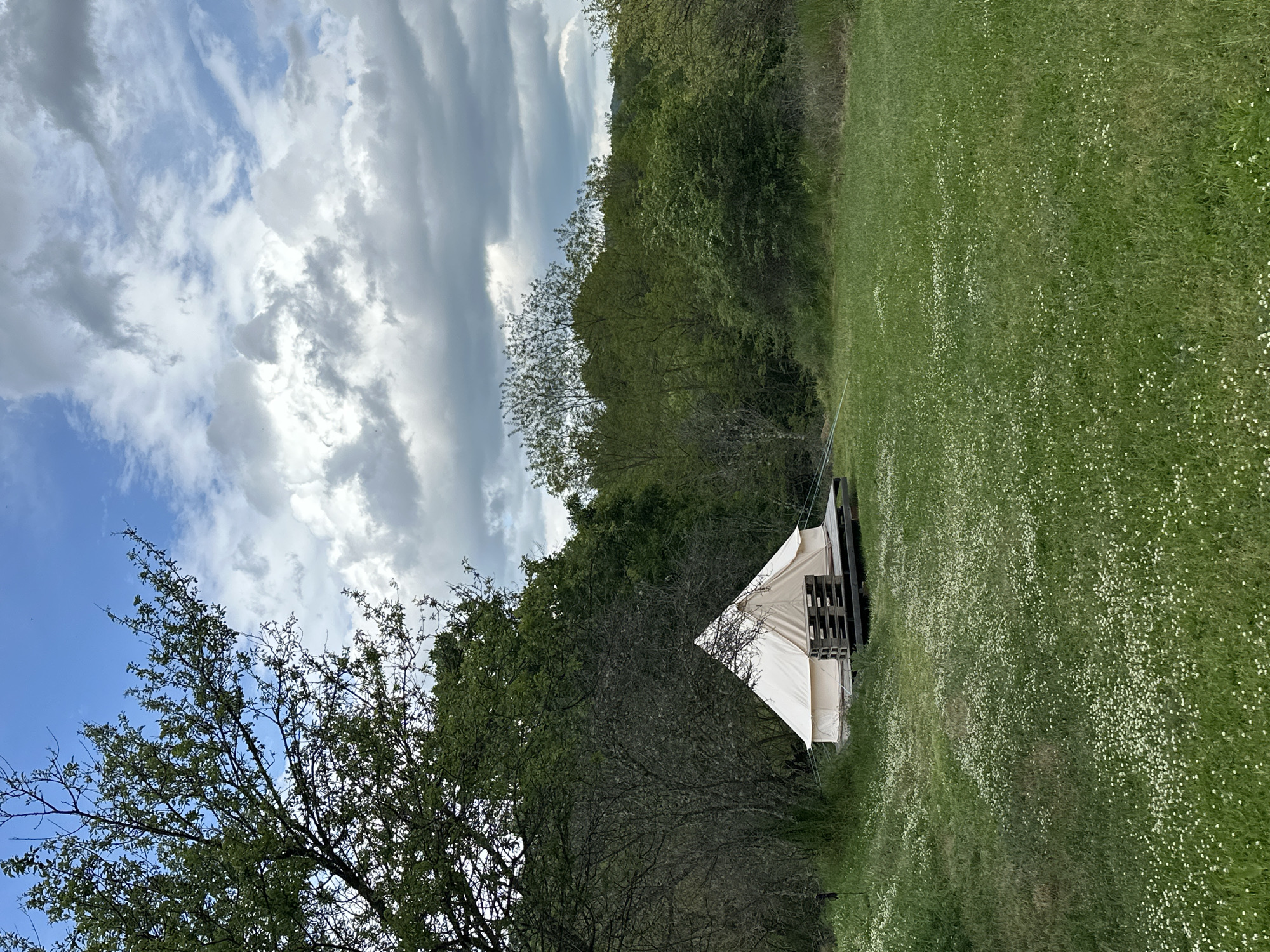A slightly rotated, medium-shot outdoor scene shows a small canvas tent set up in a grassy field next to a treeline, possibly in a rural area near Mrachenik, Bulgaria, given the location information. The tent is white and appears to be constructed on a wooden base, perhaps a makeshift platform. The field is green with many small white flowers scattered throughout. Dense trees border the field on one side. Above, the sky is partly cloudy with a mix of white and grey cloud formations against a blue background. In the lower-left, a tree with bare branches is visible. There are no people or animals apparent in the image.