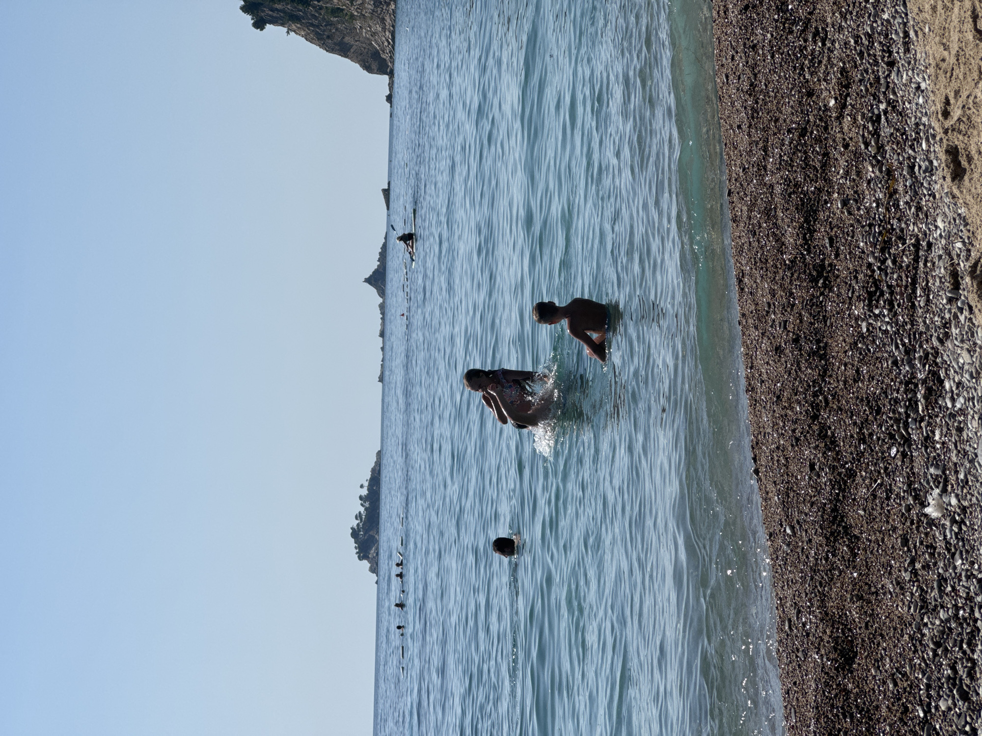 The image shows a beach scene in Đurovići, Montenegro. Several people are in the clear blue water, with some swimming and playing. In the foreground, a boy is sitting in the water, facing away from the camera. To his left, another person is partially submerged, with their head and shoulders visible. Further out in the water, a group of people are seen in the distance, with some appearing to be swimming or floating. A lone kayaker is visible on the water in the middle ground, paddling towards the right of the frame. In the background, small islands with trees are dotted across the horizon, and a rocky cliff face dominates the right side of the image. The beach itself is covered in pebbles, with a hint of sand in the lower right corner. The sky is clear and blue, indicating a sunny day.