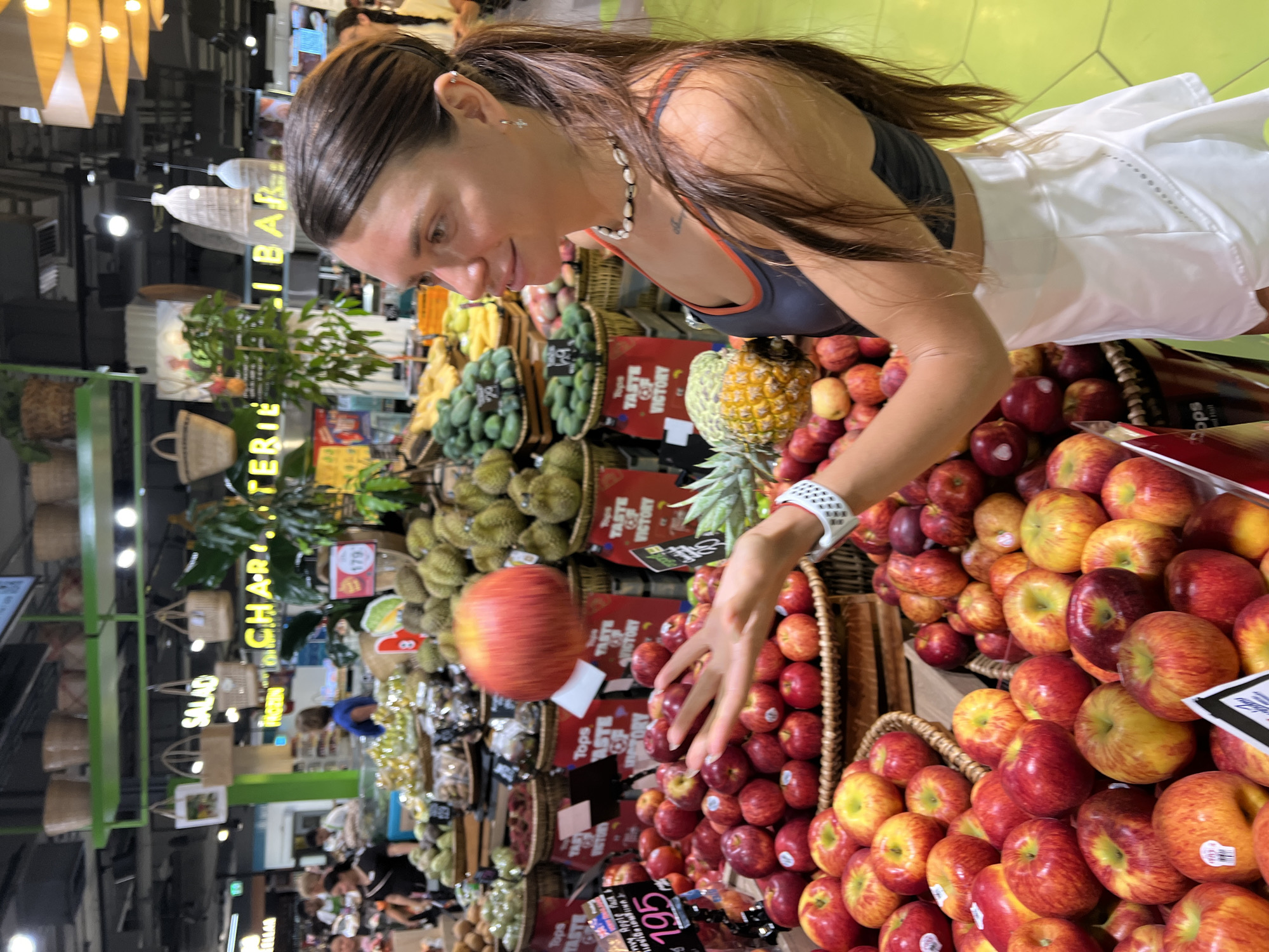 In this vertical, medium shot, a woman with long brown hair is looking down at a basket of apples. She is wearing a grey sports bra with orange trim and a white skirt. Her right arm is extended, and her hand is reaching towards the apples. There's a smartwatch on her left wrist. She has a necklace made of shells and a tattoo on her neck. The background appears to be a grocery store or market, with displays of fruits and vegetables like durians, pineapples, and more apples. There are also baskets and shelving units visible, along with bright, geometric lighting from above. Several signs with text such as "SALAD", "CHARTERIE", "Tops", "TASTE VICTORY", and prices like "195" and "179" are present, suggesting a retail environment. The overall scene suggests a casual shopping trip, with the woman actively selecting produce. The lighting is bright, typical of an indoor retail space.