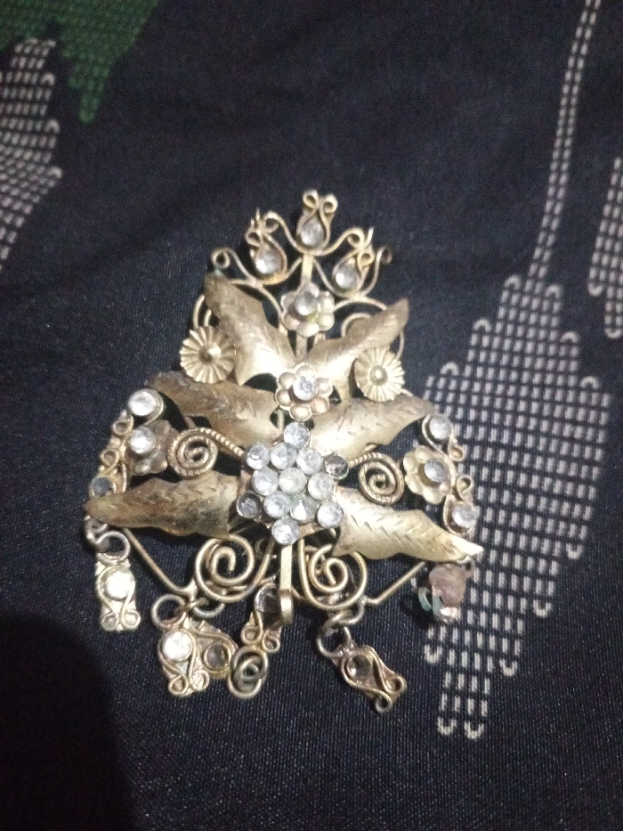 The image displays a single ornate piece of jewelry, likely a brooch or hair ornament, centrally positioned on a dark, patterned textile. The jewelry is constructed from an aged, gold-toned metal with visible tarnishing. It features a prominent central cluster of multiple small, clear, round, faceted stones. Surrounding this cluster are stylized metal leaves and smaller floral elements, each containing a single clear stone. Intricate filigree work with swirling patterns extends from the main body, incorporating additional clear stones and several small dangling components at the bottom, some of which are adorned with stones. The background is a dark fabric, possibly black or deep blue, patterned with a repeating geometric design in a lighter color, resembling a series of inverted U-shapes or vertical dashed lines. A hint of a green surface is visible in the upper-left corner. The scene is located in Hasanpur, India. No individuals, actions, or direct interactions are visible.