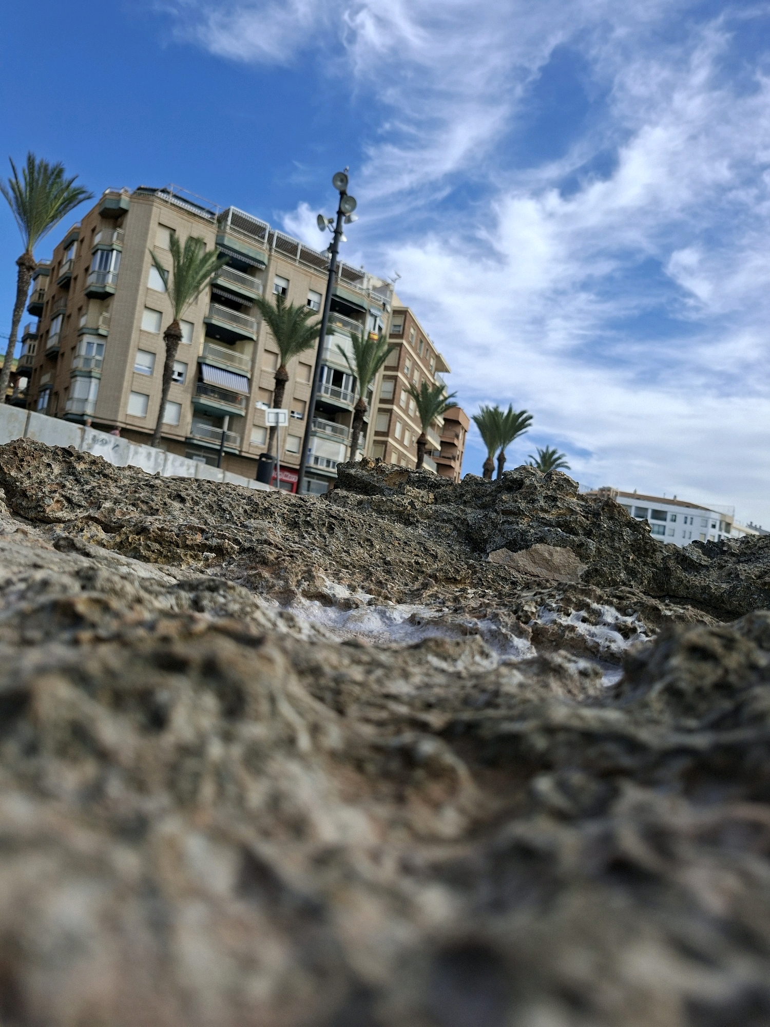 The image is a low-angle shot of a rocky foreground with buildings and palm trees in the background, under a bright blue sky with wispy clouds. In the mid-ground, there are several buildings, including a beige, multi-story apartment building with balconies and a white building further in the distance. Palm trees are visible in front of the apartment building. A street lamp with multiple lights is positioned on the right side of the frame. The foreground consists of rough, textured rocks with some white salt deposits. The overall scene suggests a coastal urban environment, possibly in Torrevieja, Spain, given the architecture and vegetation typical of the region. There are no visible people or ongoing actions in the image.