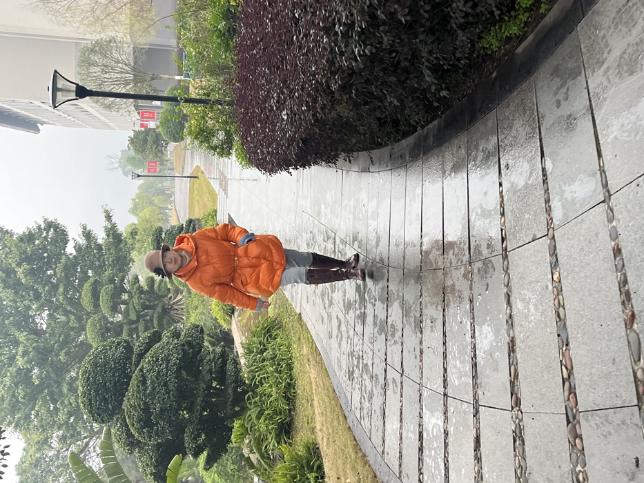 A person wearing an orange jacket, grey pants, brown boots and a tan hat walks on a wet, gray sidewalk. The sidewalk is bordered by green vegetation on one side and a dark hedge on the other. Streetlights are visible in the distance, along with buildings shrouded in mist. The scene appears to be in a park or garden.