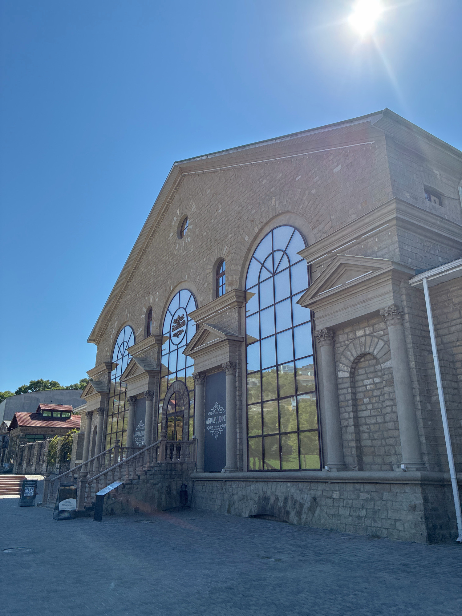 The image shows a large, ornate building with a stone facade, arched windows, and classical columns, located in the city of Abrau-Dyurso, Russia. The sun is shining brightly in a clear blue sky, creating a lens flare in the upper right portion of the image. The building appears to be a historical structure, possibly a winery or a museum, given the signage visible on the windows and entrance. In front of the building, there is a paved courtyard with some stairs leading up to the entrance. The courtyard has a few signs with QR codes, suggesting a tourist attraction. Trees and other buildings are visible in the background. The overall scene depicts a sunny day at a prominent landmark in Abrau-Dyurso.