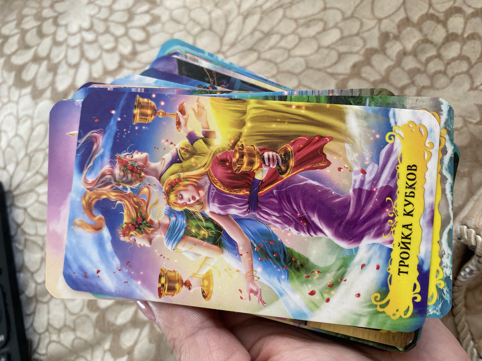The image shows a close-up of a hand holding a tarot card with an illustration of three women holding golden cups. The card has a yellow banner at the bottom with Russian text that reads "ТРОЙКА КУБКОВ" (Troika Kubkov), which translates to "Three of Cups". The background of the card depicts a vibrant sky with swirling clouds and falling rose petals, and the women are adorned with floral wreaths. A stack of other tarot cards is visible behind the main card, fanned out to the right. The hand holding the card has fingernails painted with a light pink polish. The background behind the cards is a textured, light-colored fabric. The image does not contain any information or visual cues that would indicate its location in Chornomorsk, Ukraine.