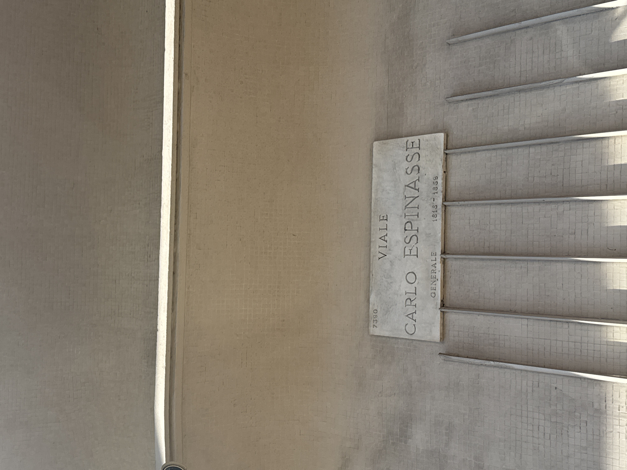 The image shows a sign in Milan, Italy, indicating "VIALE CARLO ESPINASSE GENERALE 1815-1859". The sign is a rectangular plaque mounted on a wall made of small, light-colored square tiles. To the right of the plaque are several horizontal metal bars or railings. The background wall is a light beige color, and there's a section on the left side with a vertical line that appears to be a structural element. The lighting suggests an outdoor environment. The number "7390" is visible on the plaque, possibly a street address.