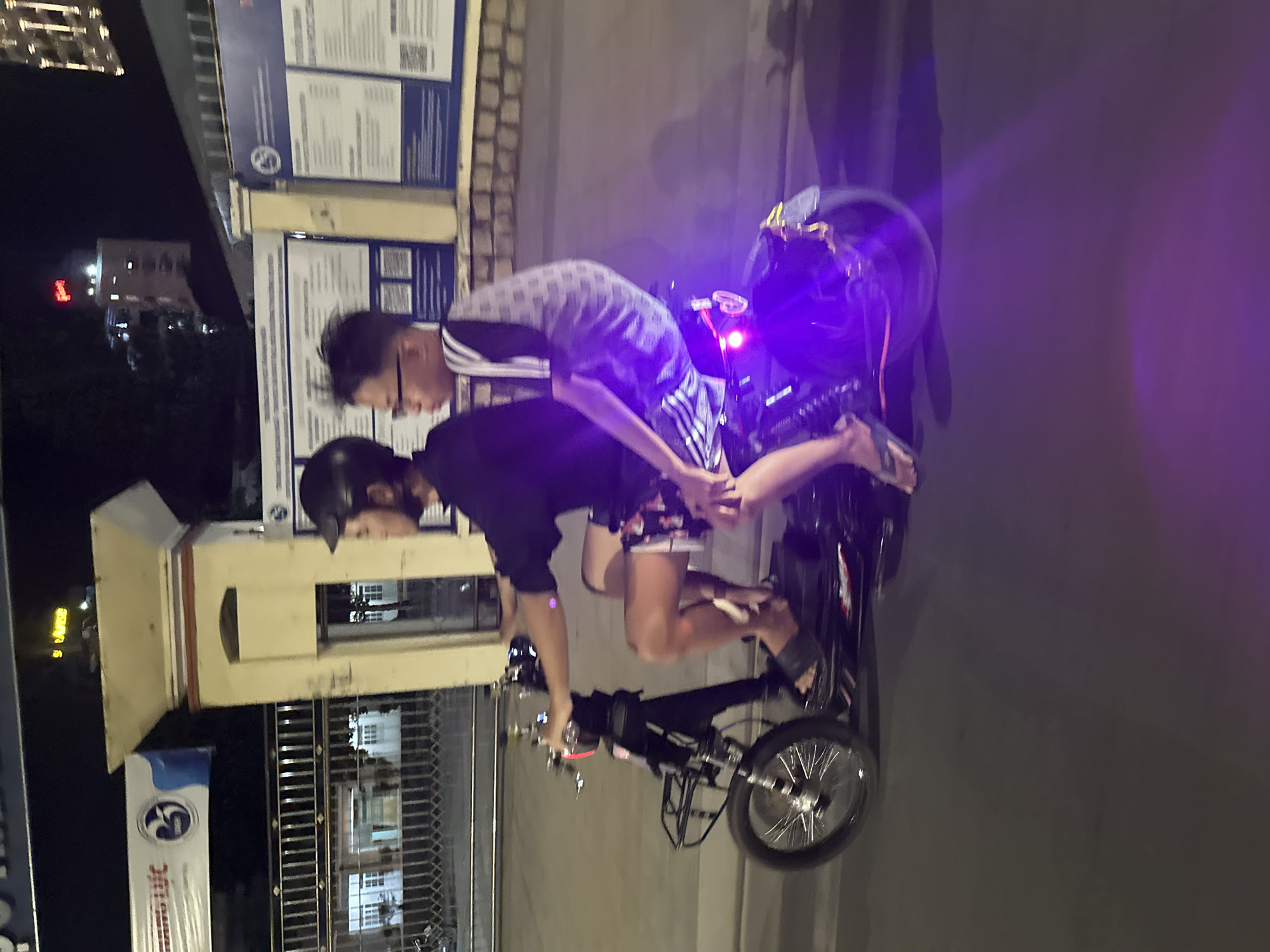 Two individuals are riding on an electric bike at night in what appears to be the Bac Nha Trang Ward, Vietnam. One person is wearing a helmet and a black shirt and the other is wearing eyeglasses and a striped shirt. A streetlight illuminates a building nearby, and signs are attached to it.