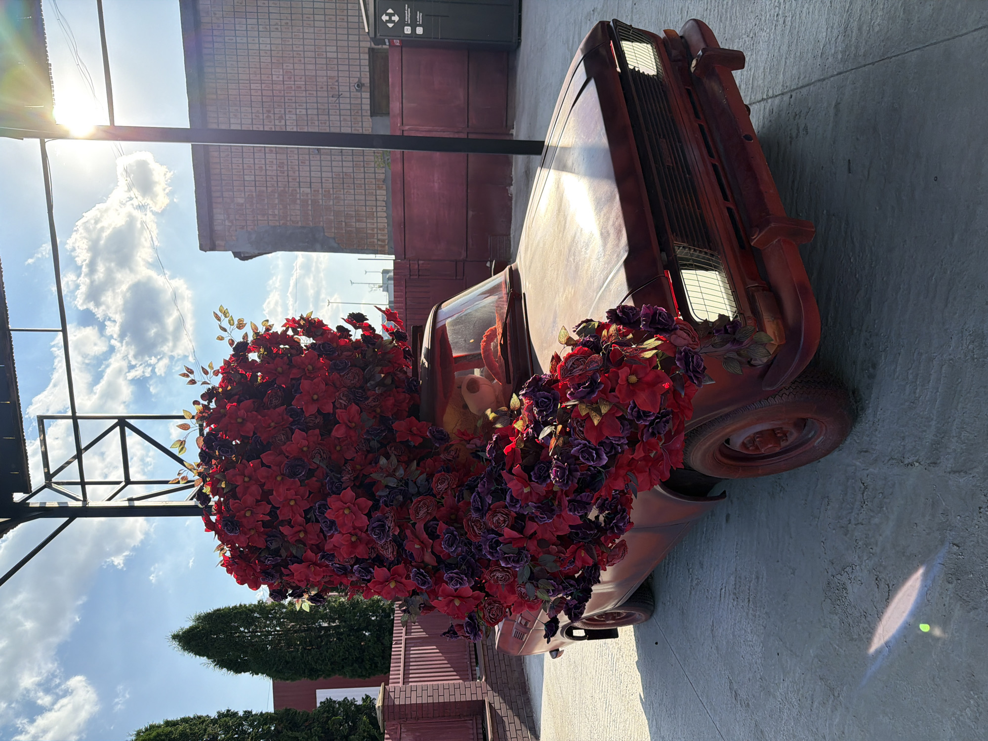 The image depicts a vintage red car, heavily decorated with red and purple artificial flowers, parked outdoors. The car appears to be a Moskvitch 2140. The flowers are arranged in a chaotic manner, spilling over the hood, roof, and front grille. A portion of the interior is visible, showing a light-colored dashboard and a hint of a person's head, possibly a mannequin, with a blonde wig.
The car is positioned on a concrete surface, with some lens flare from the sun visible on the ground. In the background, there is a red brick building with a tiled facade, a red wooden fence, and several tall green trees. A portion of a black canopy structure is visible overhead, along with wires and a segment of a blue sky with scattered clouds. The overall impression is that of an artistic installation or a display.
The provided context states this scene is located in the city of Ternopil, Ukraine.