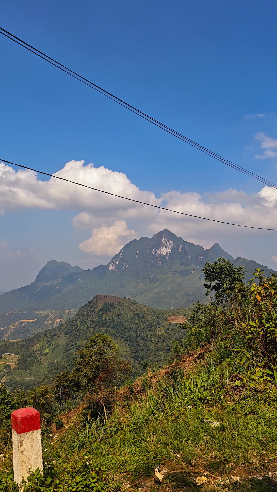 The image depicts a landscape in Mậu Duệ Commune, Vietnam, featuring mountains, a partially cloudy blue sky, vegetation, and infrastructure. The focal point is a range of mountains in the background, with the largest peak exhibiting exposed rock faces on its slopes. Green, forested vegetation covers the lower portions of the mountains. In the foreground, a roadside embankment is covered with lush green grasses and various plants. A red and white concrete post stands on the embankment's edge. Two power lines run diagonally across the upper portion of the sky. The sky is mostly blue with scattered white cumulus clouds. Patches of land with terraced agriculture are visible in the mid-ground, indicating human modification of the landscape. The scene captures a combination of natural mountainous terrain with elements of human intervention, such as power lines and agricultural land use.