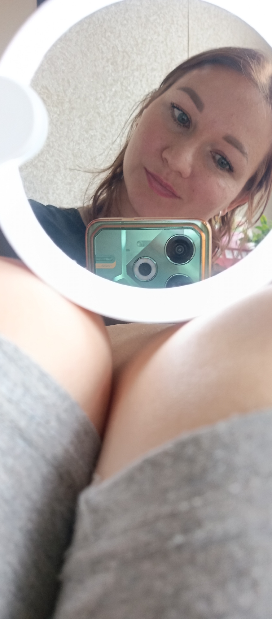 The image displays a unique selfie taken from a low angle, reflecting a person's face and a smartphone within a circular mirror or ring light. The primary subject, a person (likely female), has light brown hair, light eyes, and a neutral to slightly smiling expression, reflected in the circular object. She appears to be wearing a black top.
In the immediate foreground, the person's bare legs or upper thighs are visible, partially covered by a soft, grey fabric, possibly shorts or underwear.
Central to the reflection within the circular mirror is a smartphone, which the person is holding to capture the image. The phone is encased in a distinct light green or teal protective cover with a metallic bronze-colored frame. Its rear camera module, featuring multiple lenses (at least three visible, one with a clear ring around it), is clearly discernible.
The background of the reflection shows a light-colored, textured wall or surface. Faint hints of pink or purple objects, possibly decorative flowers, are visible in the far right of the background within the reflection. The overall scene suggests an indoor setting, and the activity is a self-portrait taken using a mirror for an interesting perspective. There is no visible text in the image.