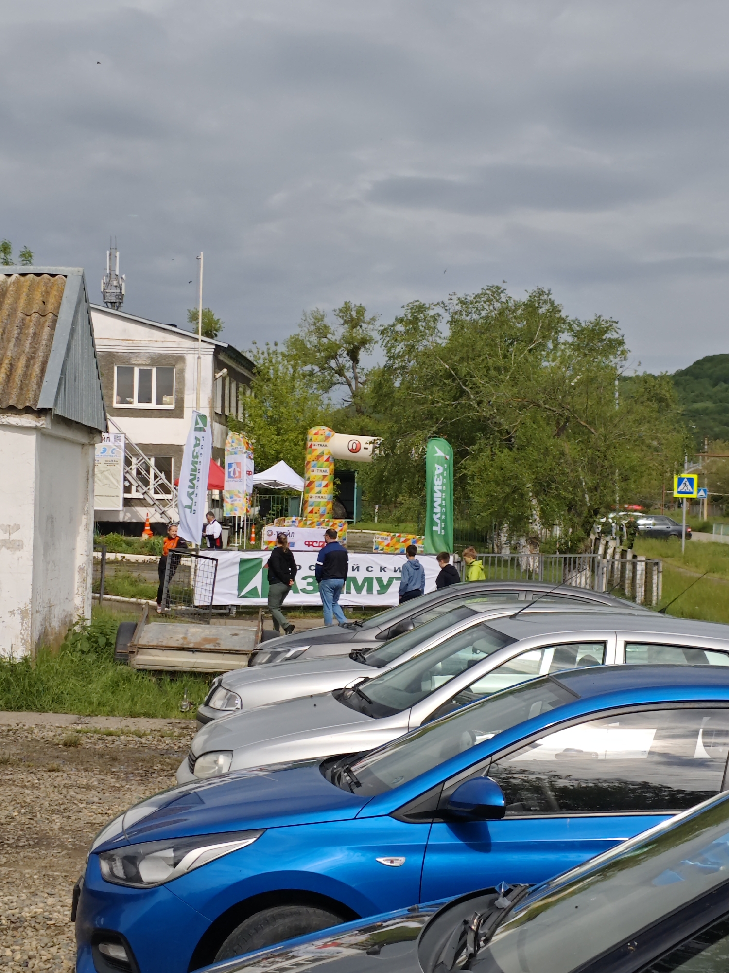 The image shows a parking area in Новоекатериновская, Russia, with a row of cars parked in the foreground. A blue car is prominent in the front row, followed by several silver cars. Behind the cars is a group of people standing near event banners and a starting gate. A two-story building is visible in the background, along with some trees and overcast sky. The environment suggests a gathering or event is taking place.