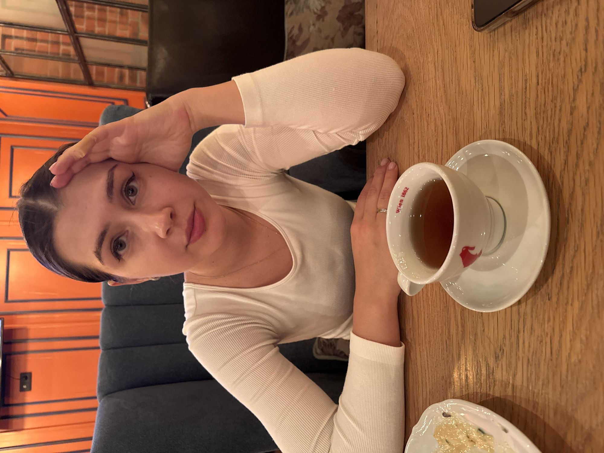 A woman is sitting at a wooden table in what appears to be a cafe or restaurant. She is wearing a white long-sleeved top and has dark hair pulled back from her face. Her left hand is resting on her forehead, and her right hand is near a cup of tea. The cup is white with a saucer and has a design on it, including the words "Wien 1862". A plate with some food on it is also visible on the table. In the background, there is orange paneling and a blue cushioned seat or booth. Based on the information available, it is not possible to determine the exact location in Krasnoyarsk, Russia.