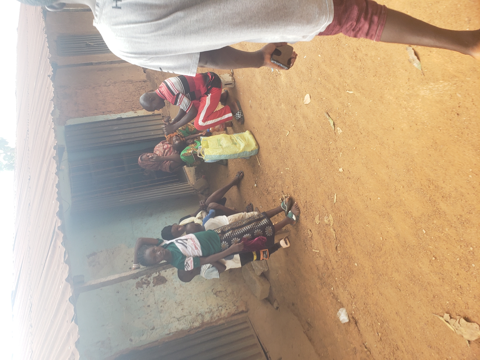 In Kachia, Nigeria, a group of individuals are gathered outside a structure with a corrugated metal roof and mud walls. In the foreground, a person wearing a grey t-shirt and maroon shorts is seen from the back, holding a mobile phone. To their left, a young girl in a green t-shirt and patterned skirt stands, holding a cloth. Behind her, two young boys and a woman are partially visible. Further back, a man in a striped shirt and red pants is seated, and another woman in a headscarf is also present. A yellow plastic bag and a water bottle are on the ground near the seated man. The environment appears to be a dusty, outdoor area.