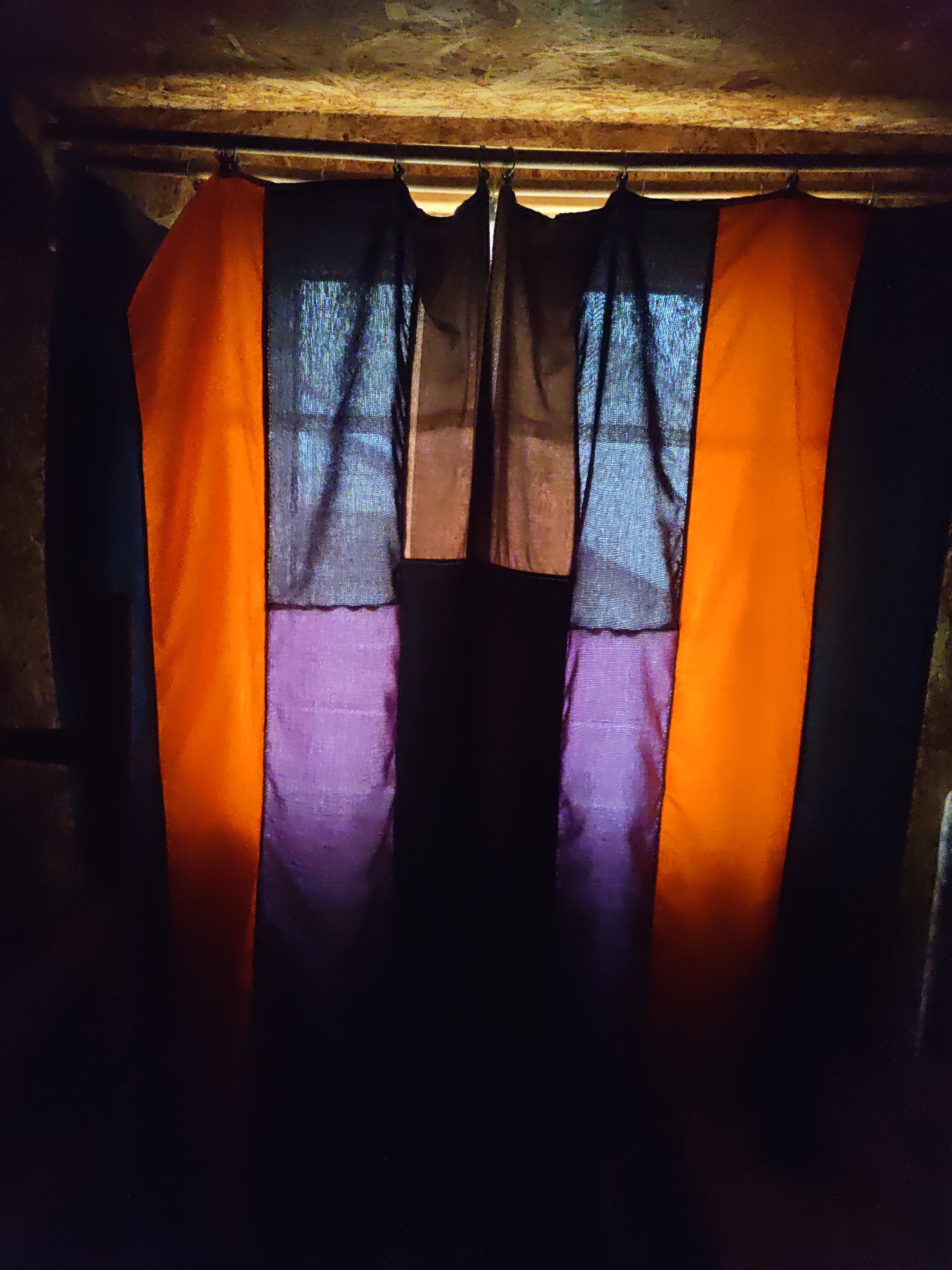 A close-up shot features a dark, rustic ceiling made of wooden planks with some light shining through gaps. Below this ceiling, a metal rod holds up a series of curtains. The curtains are made of fabric and are arranged in a pattern of vertical stripes. On the far left and far right are panels of bright orange fabric. In the center, there are panels of dark purple fabric with a textured appearance, layered over what appears to be sheer black fabric. The overall lighting is dim, with the orange panels catching the most light, casting a warm glow. The environment suggests an indoor setting, possibly a storage area or a rustic room, with no people or identifiable objects beyond the curtains and the ceiling structure. The location of Platoshino, Russia, is not evident from the image itself.